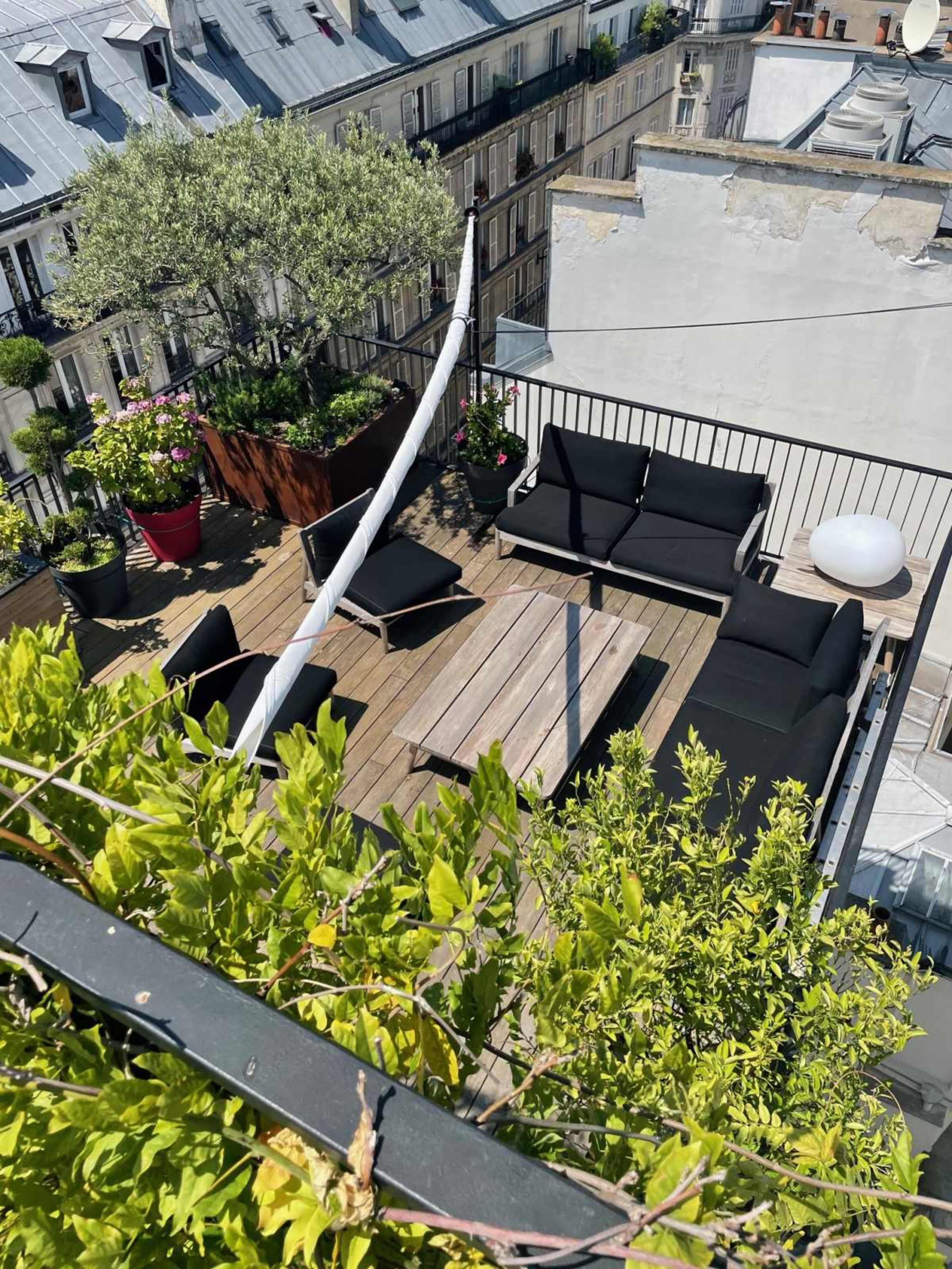 The image shows a rooftop terrace with wooden decking, black seating, a wooden table, potted plants, and a view of adjacent buildings.