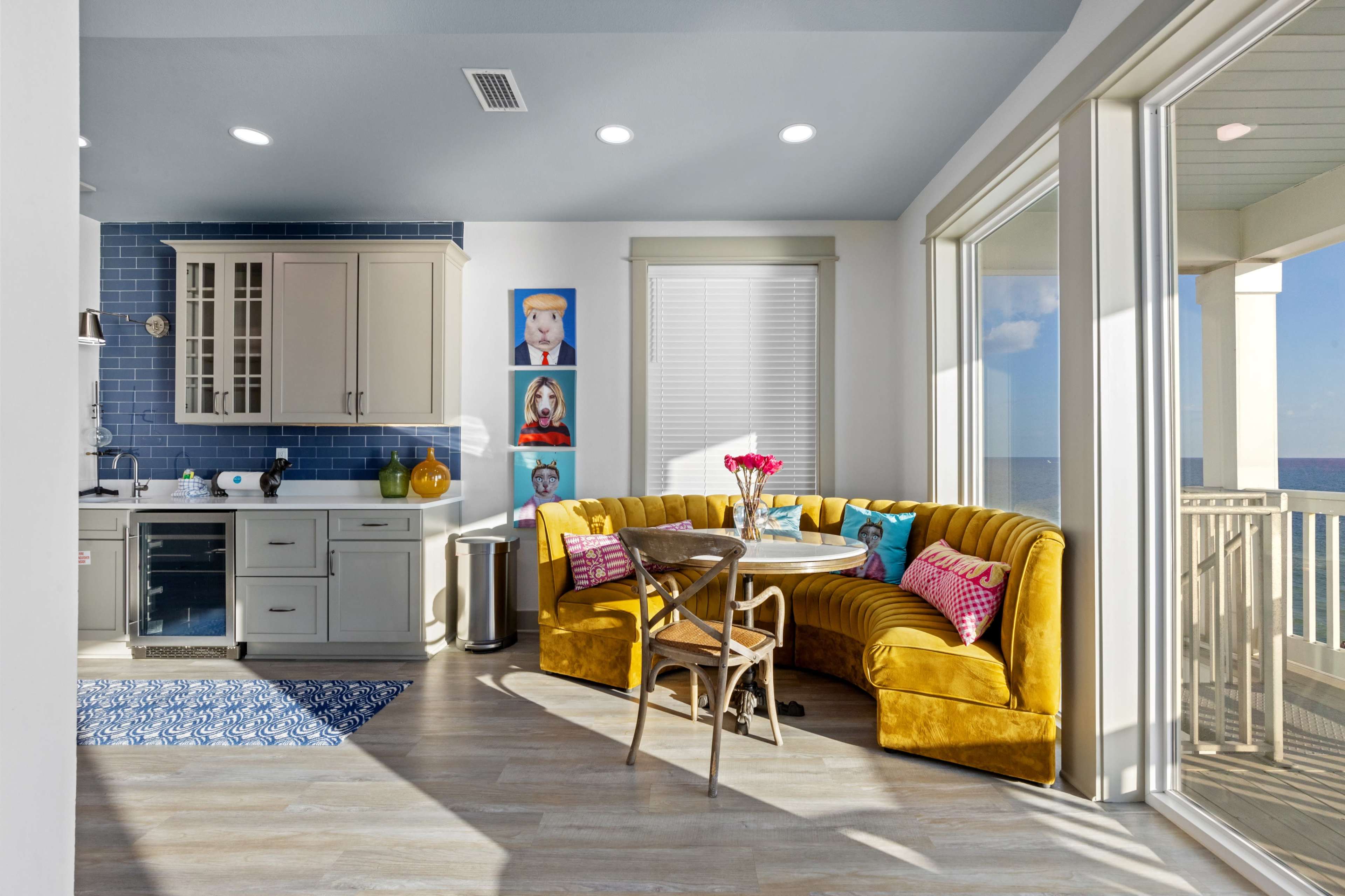 A bright kitchen area features a yellow curved banquette, a round table, and blue tiled accents, with large windows showcasing a view of the water.
