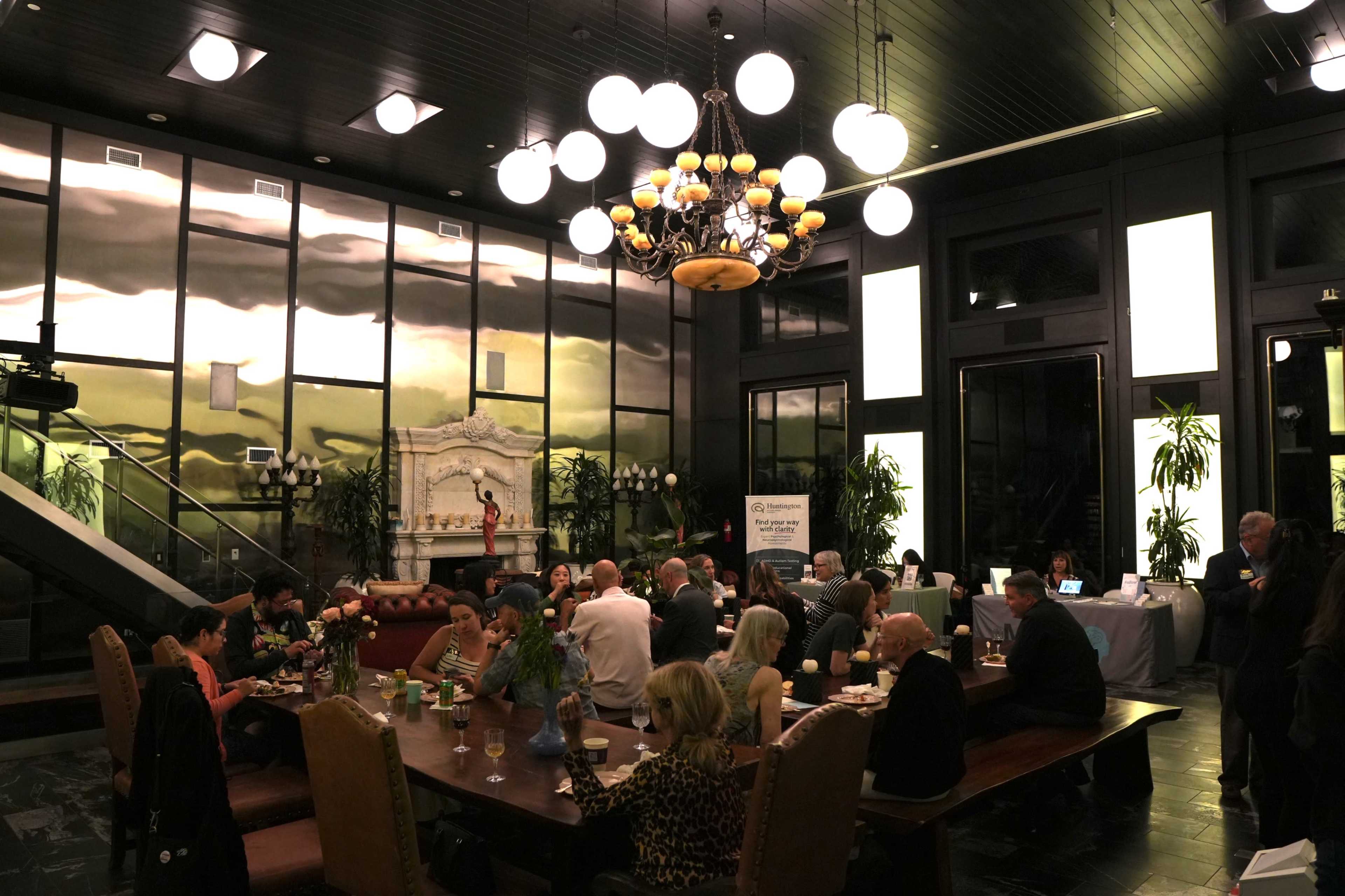 A group of people sits at a long table in a dimly lit room with large windows, plants, and a chandelier overhead.