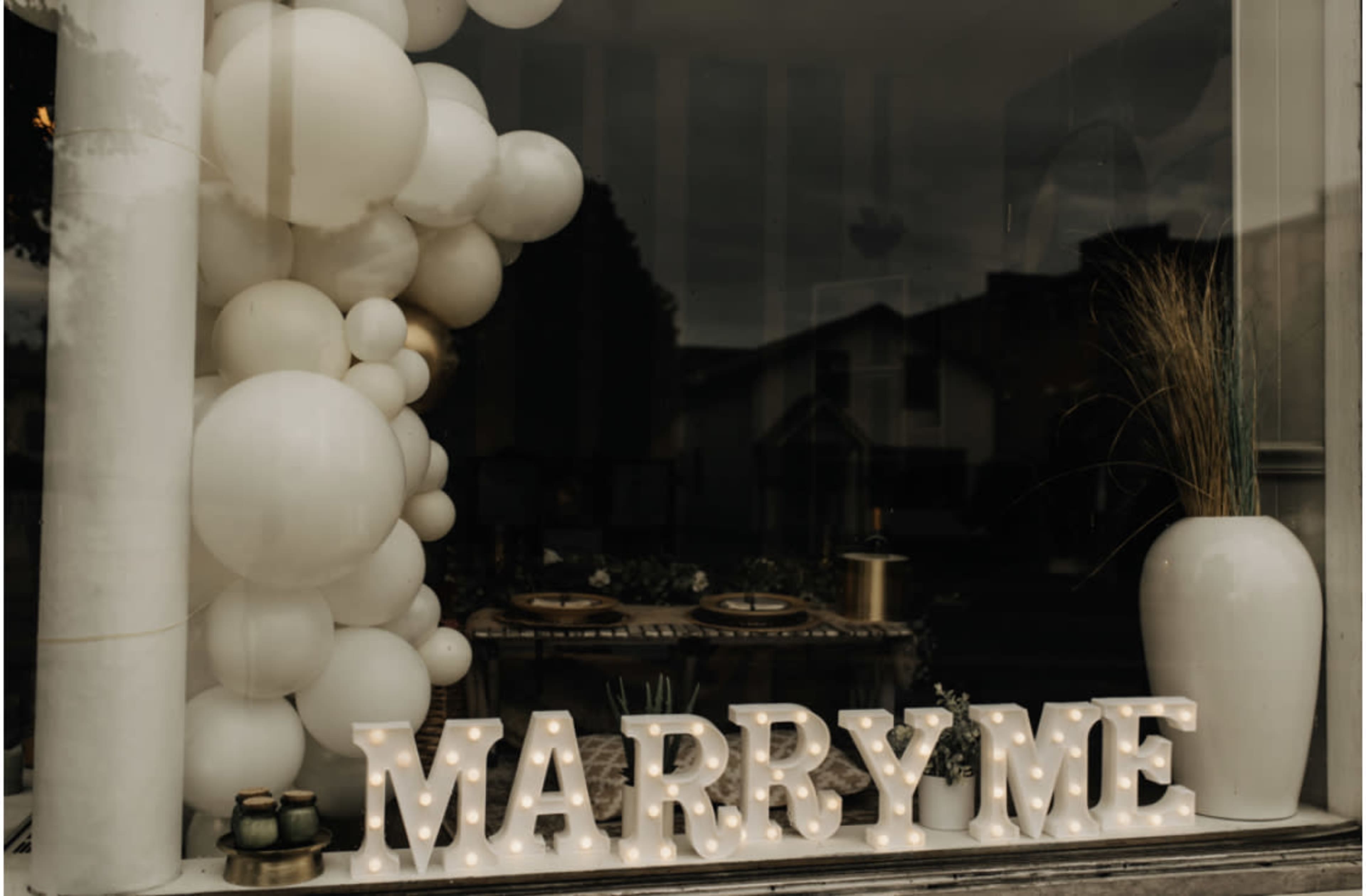 A window display features a "MARRY ME" sign illuminated with lights, accompanied by a cluster of white balloons and decorative elements.