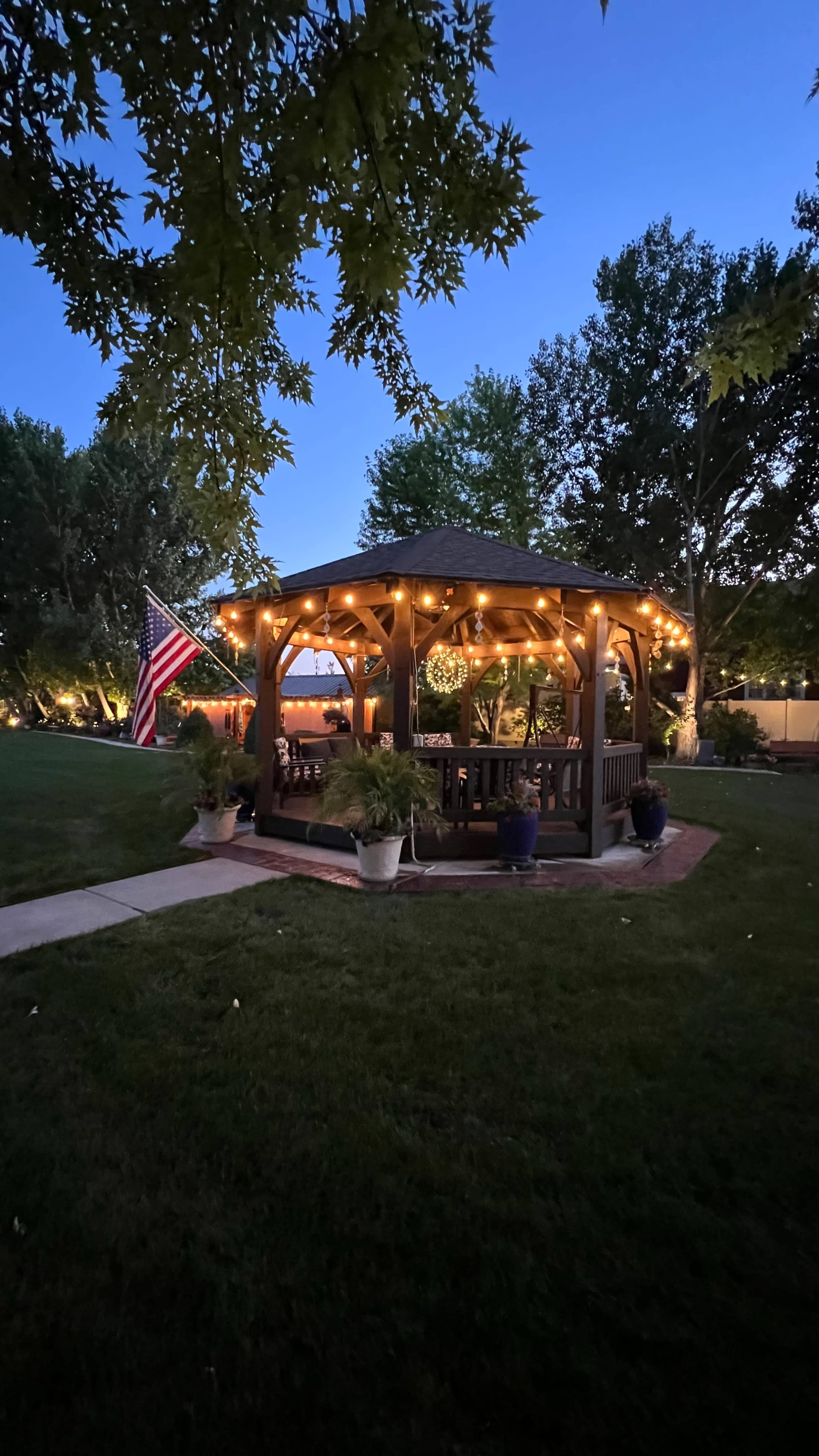 A wooden gazebo adorned with string lights stands in a well-maintained yard, with an American flag displayed nearby under a twilight sky.