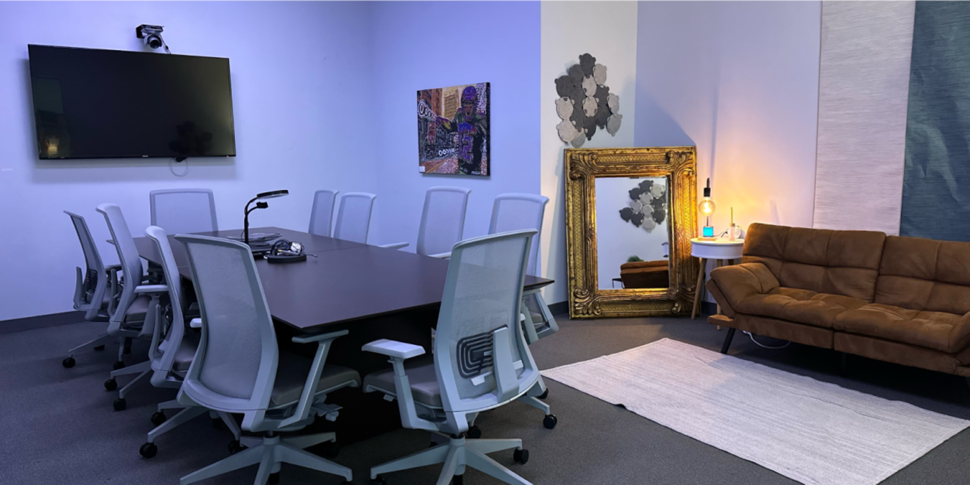 A modern conference room featuring a long table surrounded by chairs, a large screen on the wall, a decorative mirror, and a brown couch.