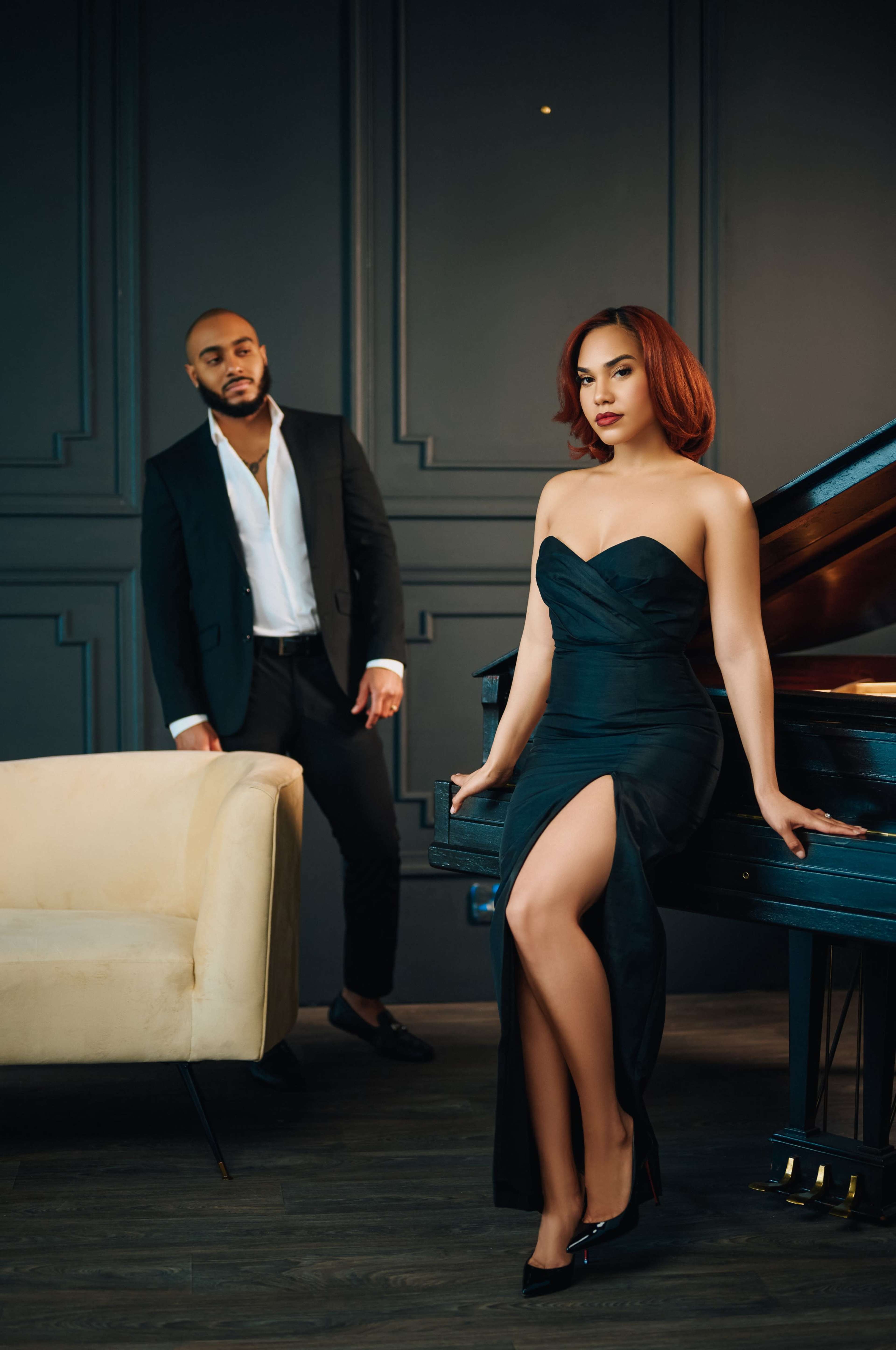 A woman in a black dress poses by a grand piano while a man in a suit stands nearby.