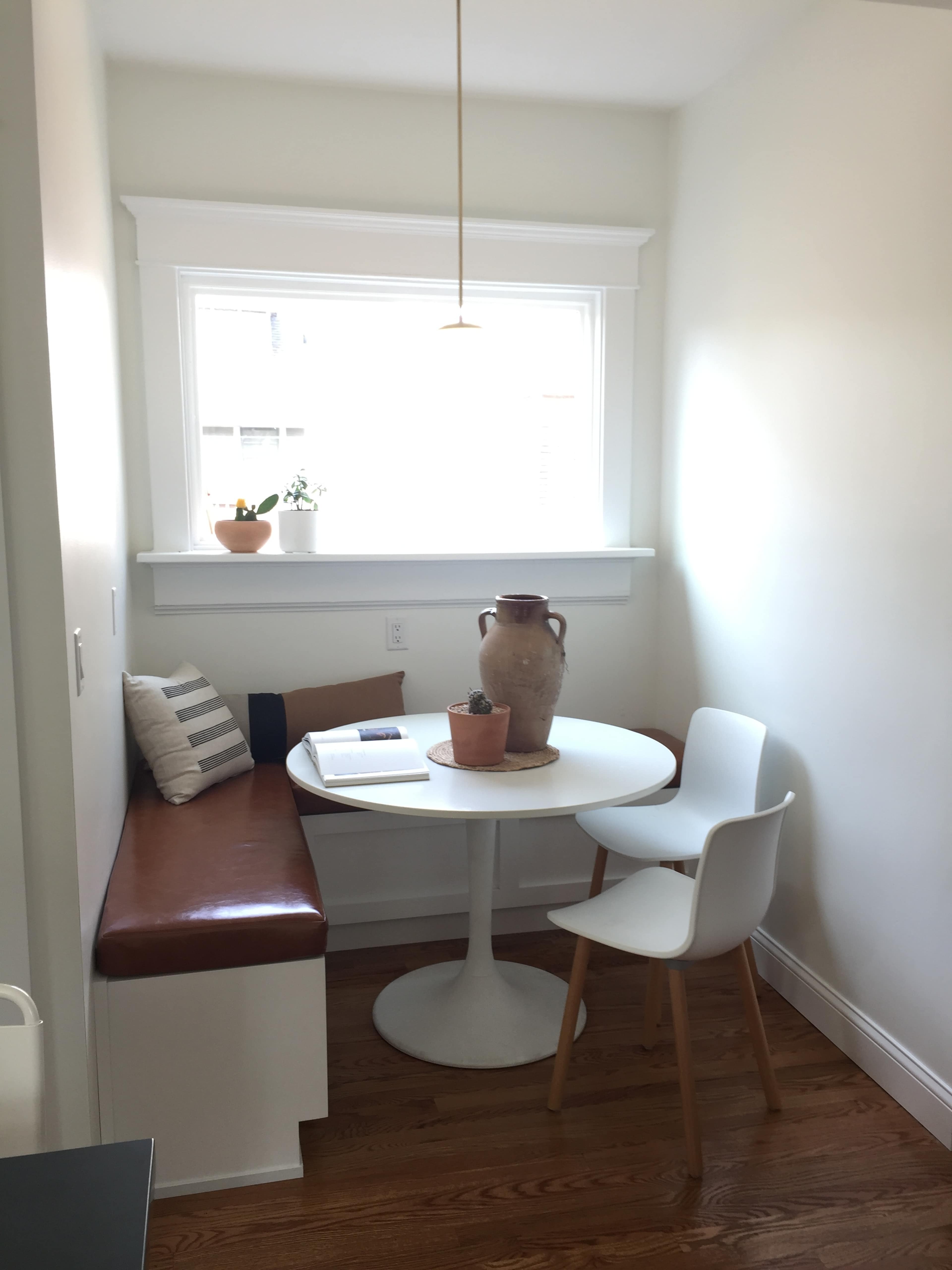 A small, well-lit dining nook features a round white table surrounded by two chairs and a built-in leather bench with decorative pillows.