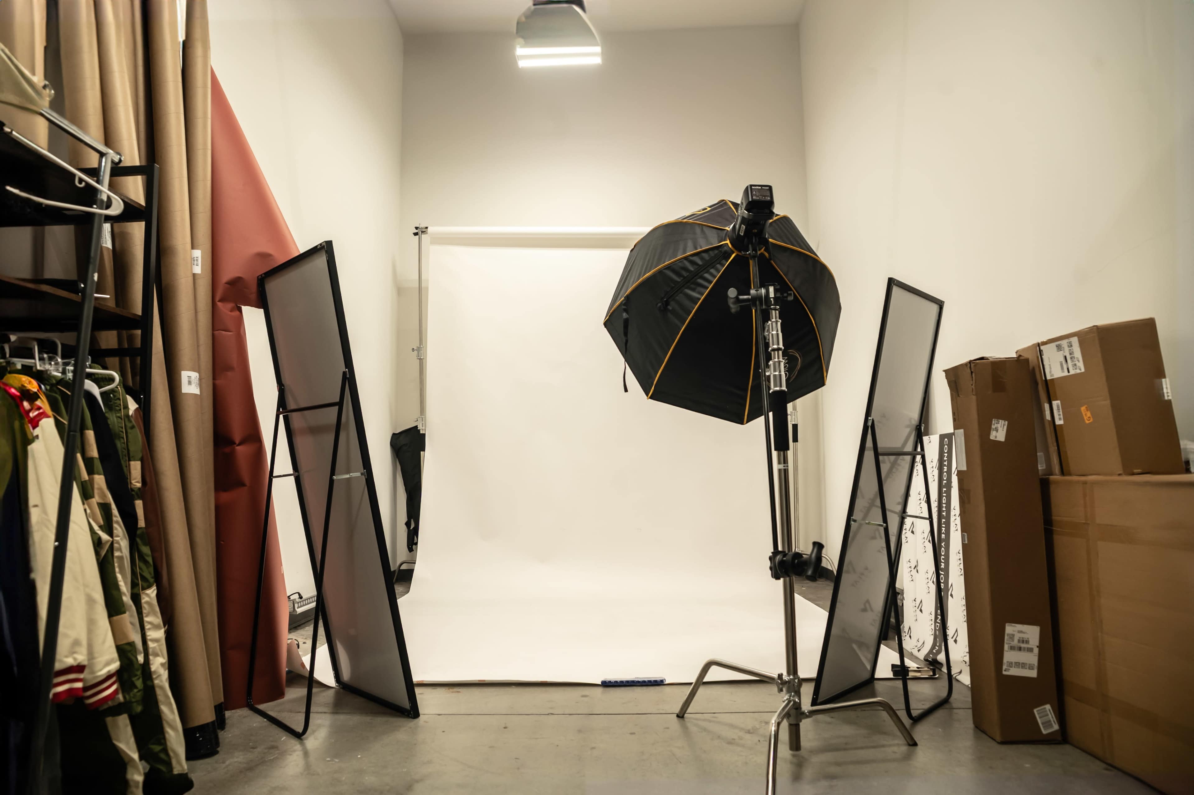 A photography studio setup features a white backdrop, softbox light, and various clothing items on racks nearby.