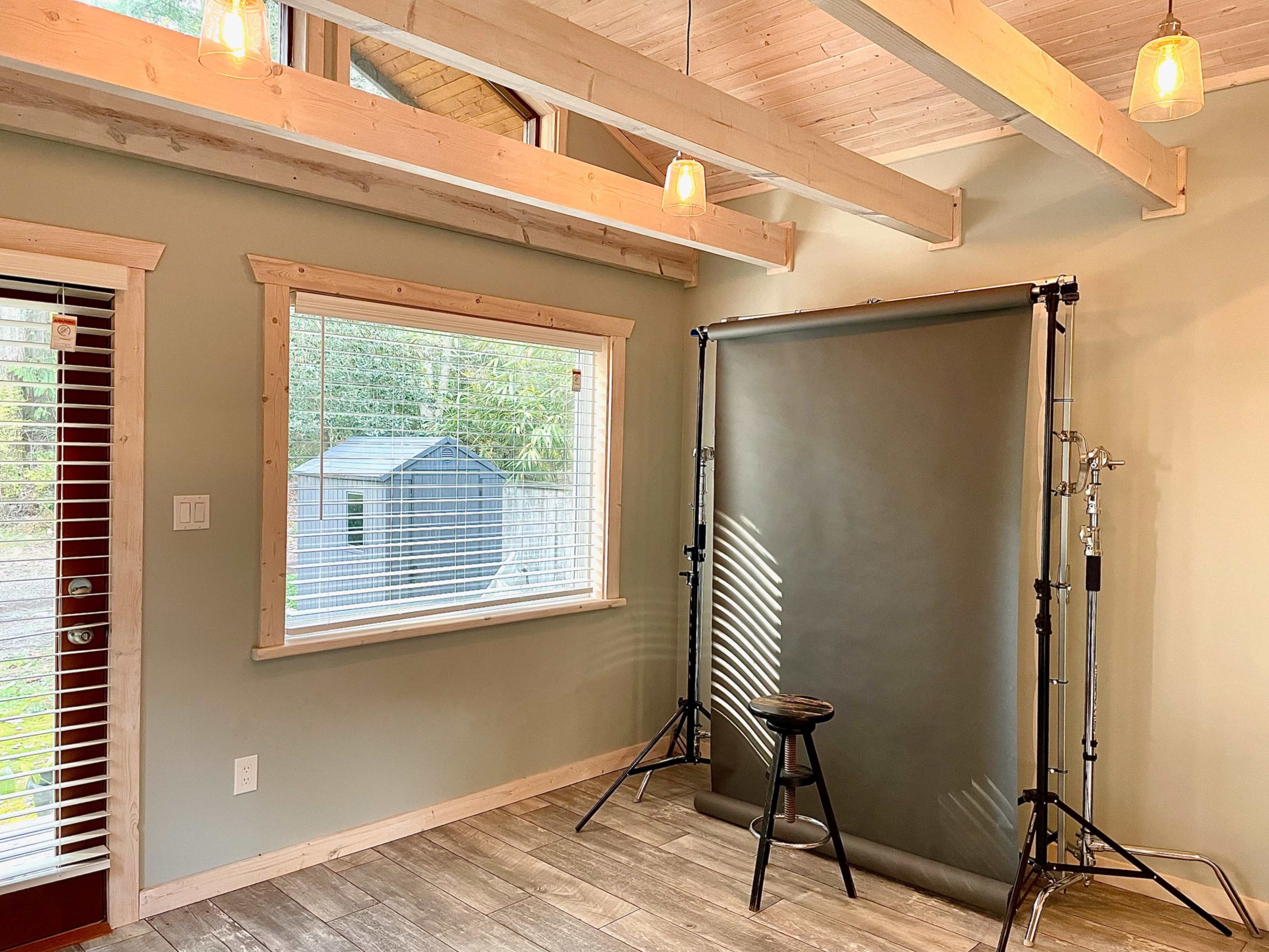 The image shows a well-lit room with wooden beams, featuring a backdrop set up for photography and a stool positioned in front of it.