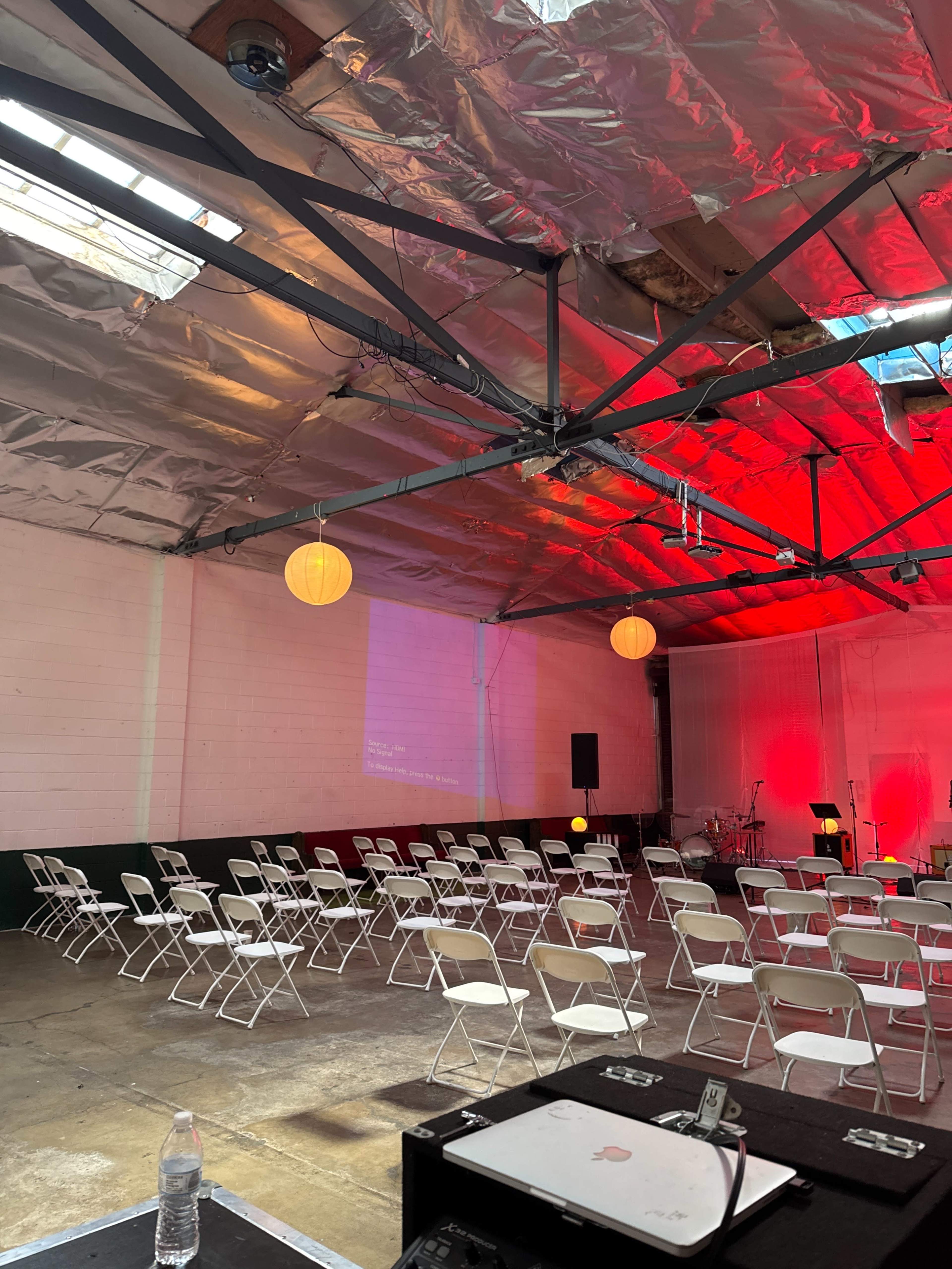 The image shows an indoor event space with rows of white folding chairs arranged in front of a stage, illuminated by red and yellow lights.