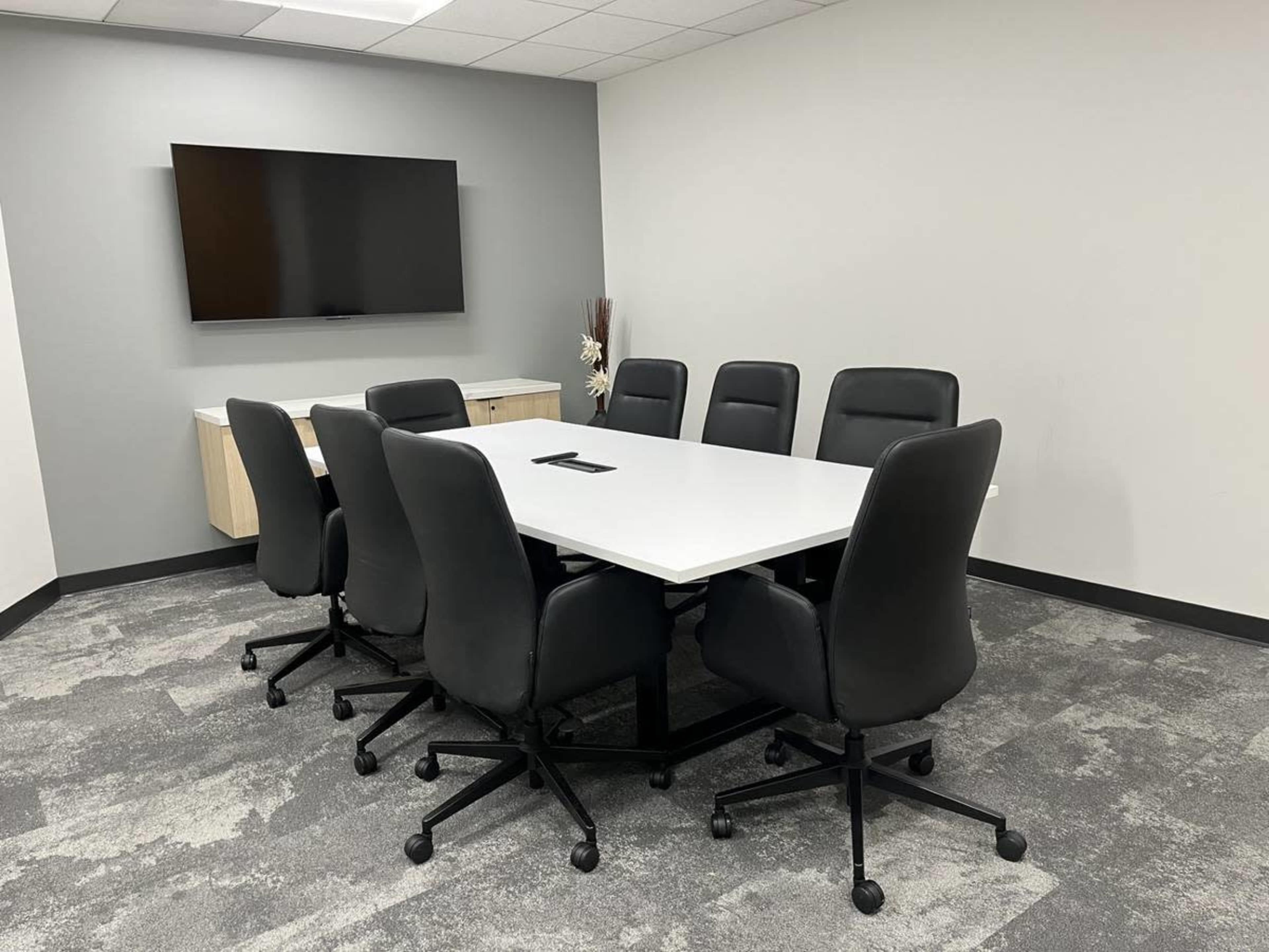 A conference room is set up with a large white table surrounded by six black rolling chairs, and a wall-mounted TV is visible in the background.