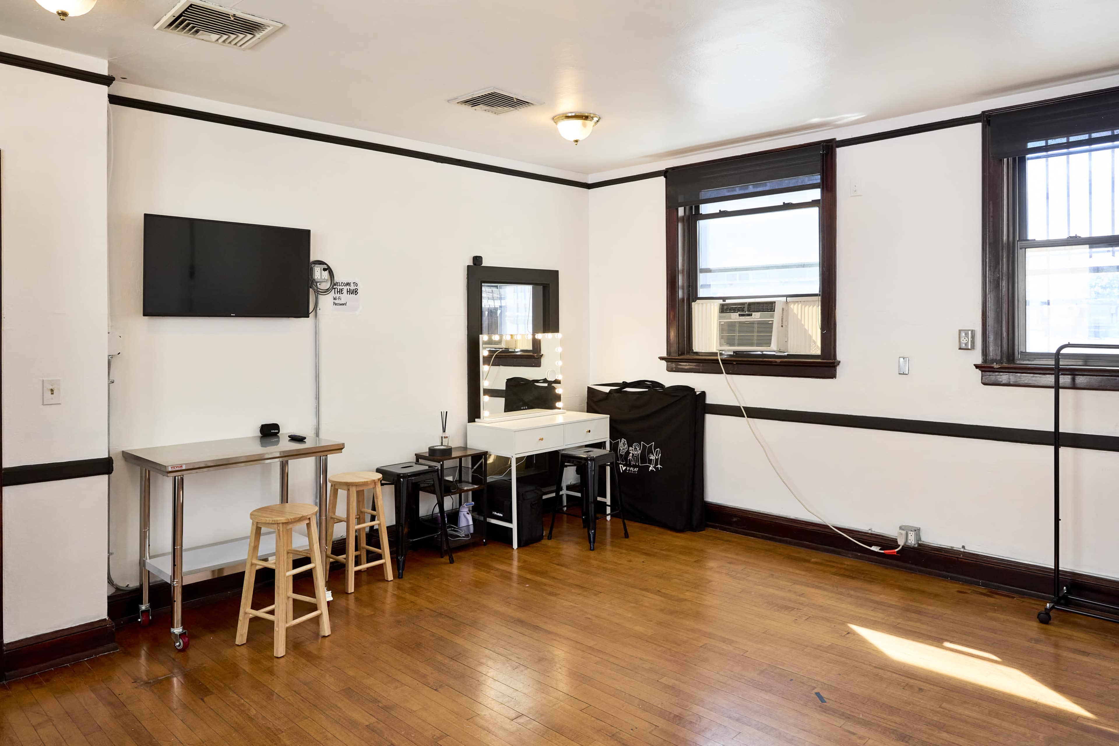 The image shows a small, well-lit room with wooden floors, featuring a wall-mounted TV, a mirror on a vanity, and various furnishings including a folding table and stools.