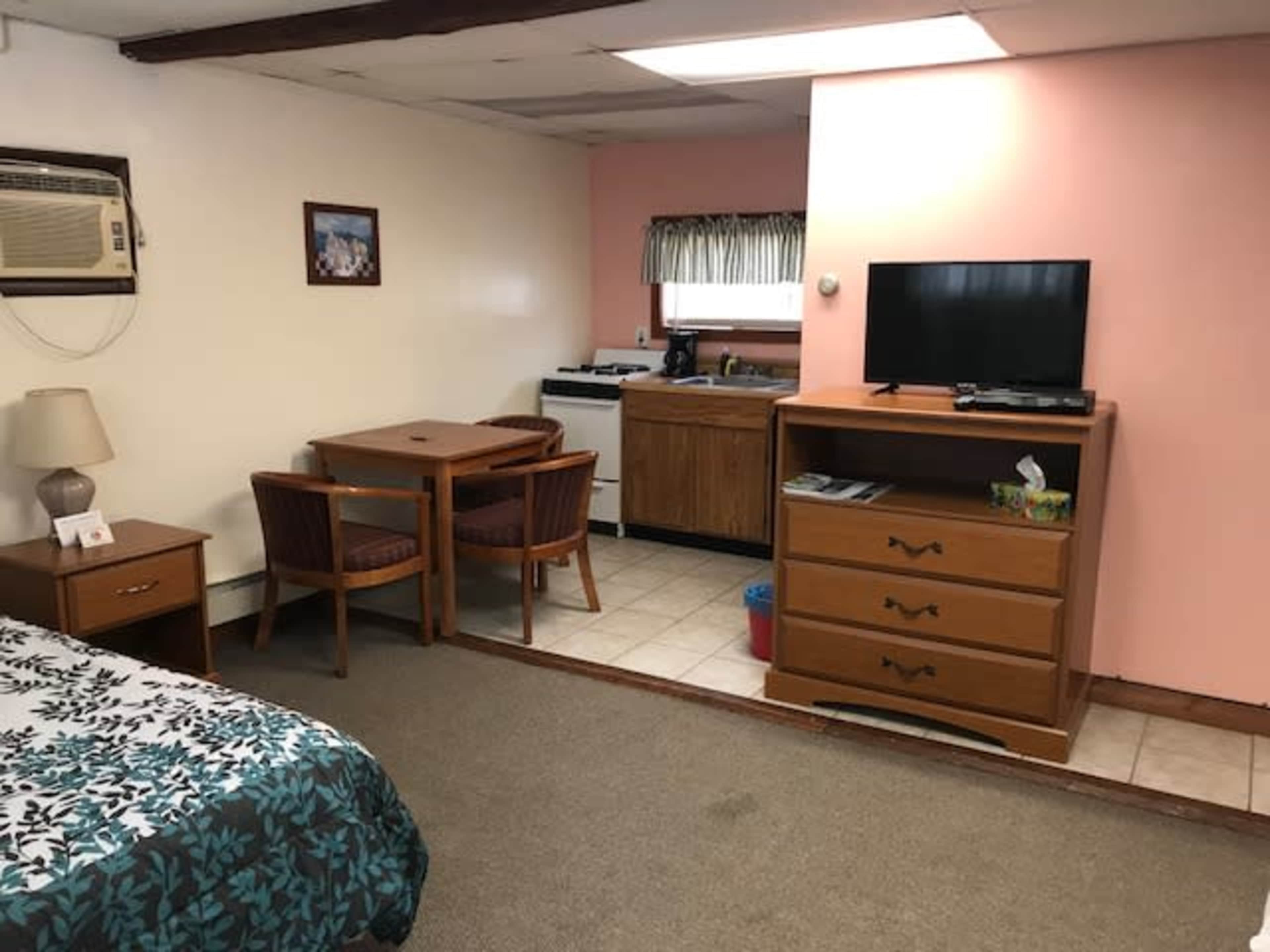 A small hotel room with a bed, a dining table, a kitchen area, a television, and a wooden dresser.