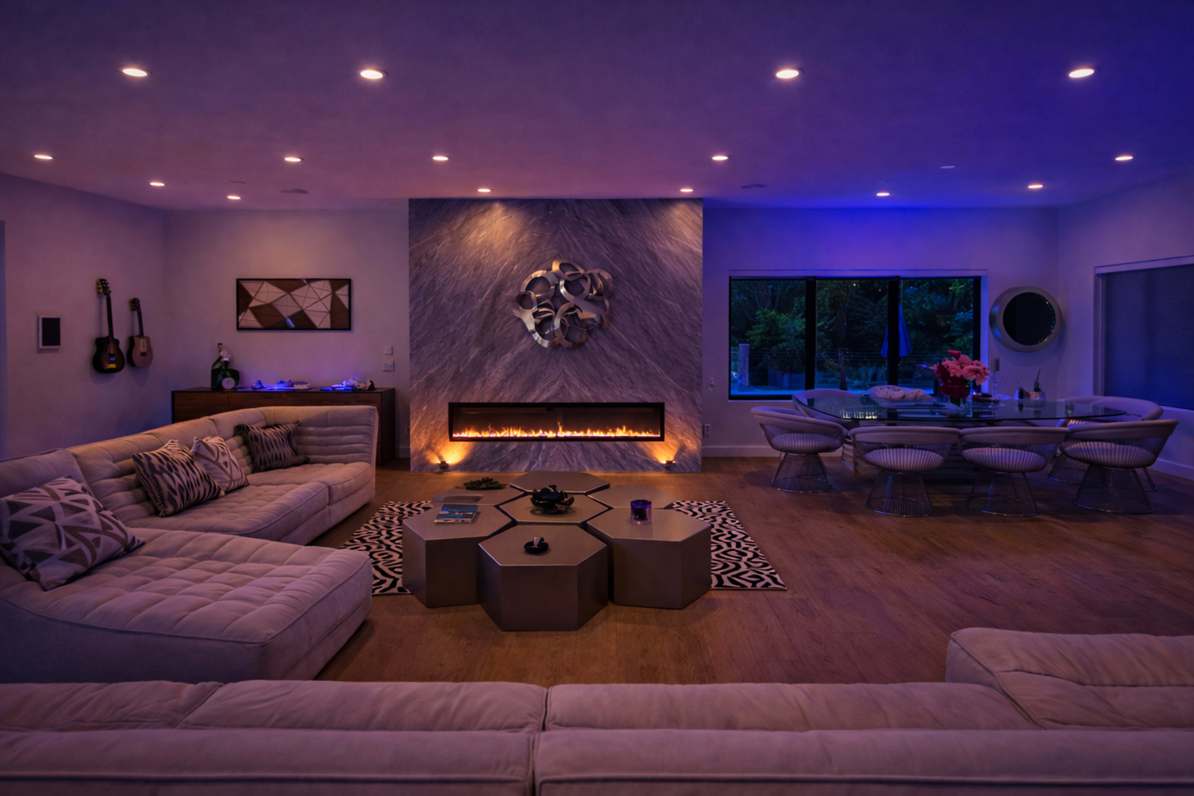 The image shows a modern living room with ambient lighting, featuring a sectional sofa, a fireplace, and a dining area with a glass table.
