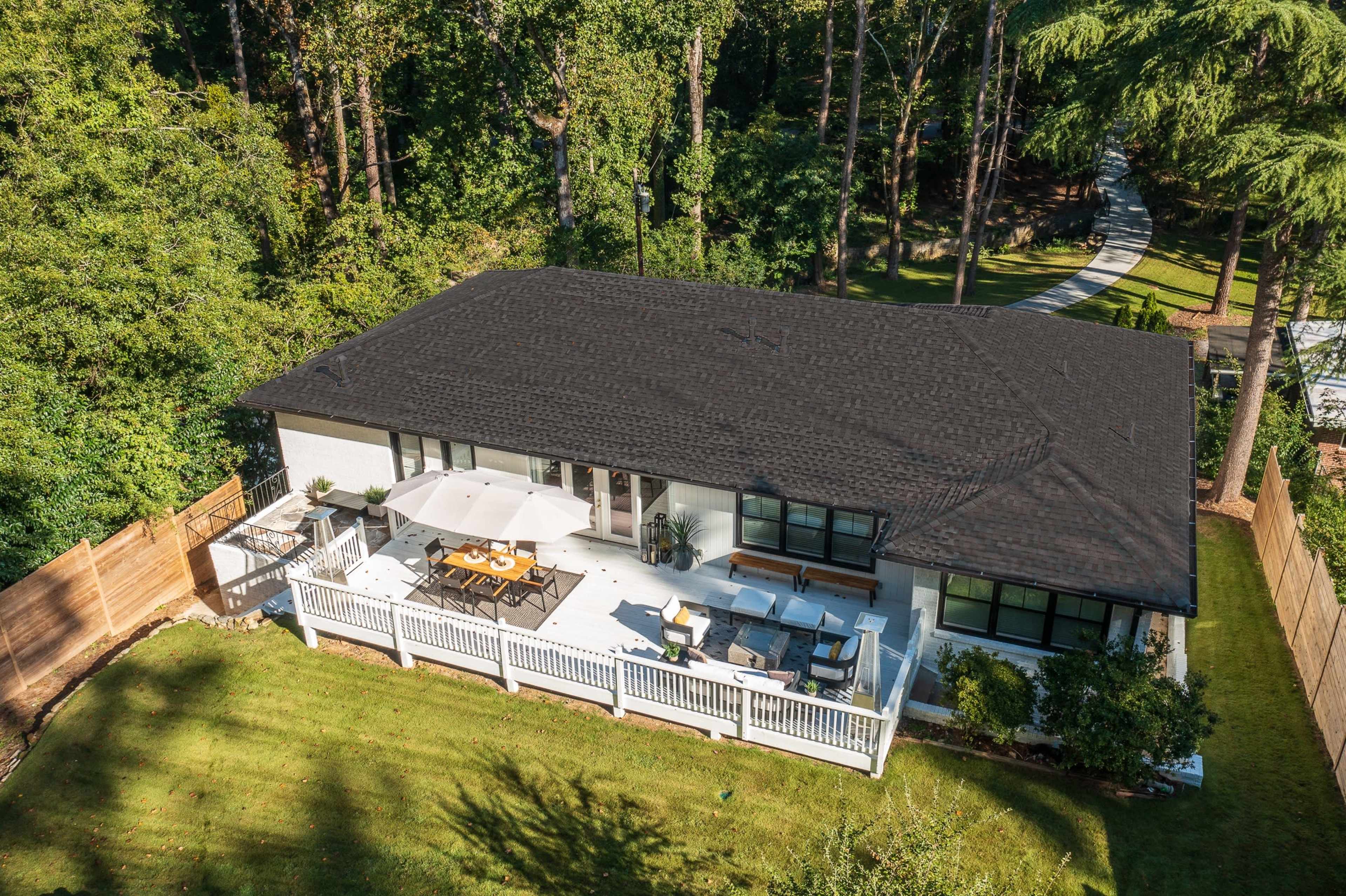 A modern single-story house features a spacious deck with outdoor seating and a dining area, surrounded by trees and a wooden fence.