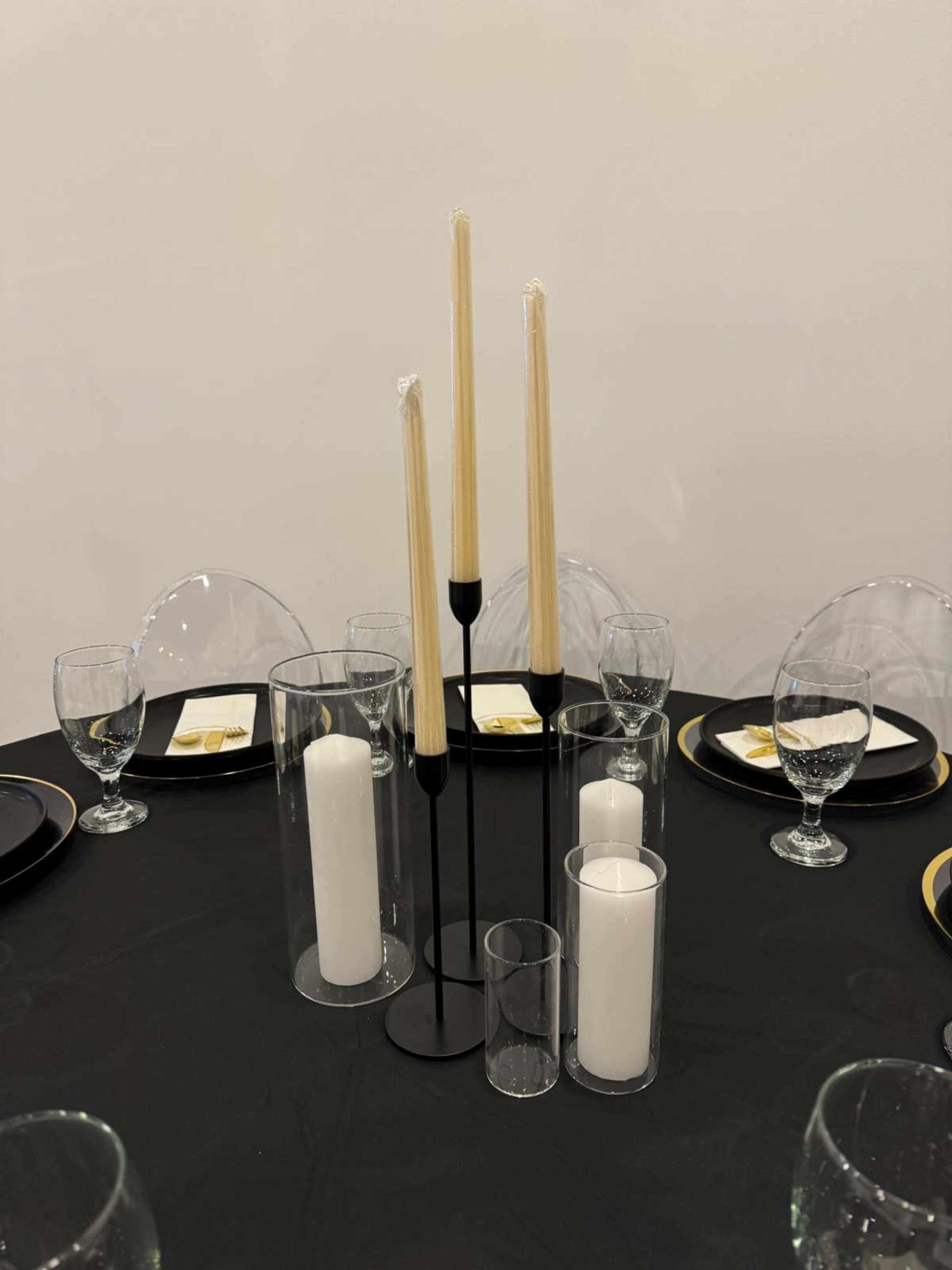 A black tablecloth is set with multiple glass plates, cutlery, and several candles arranged in glass holders at the center.