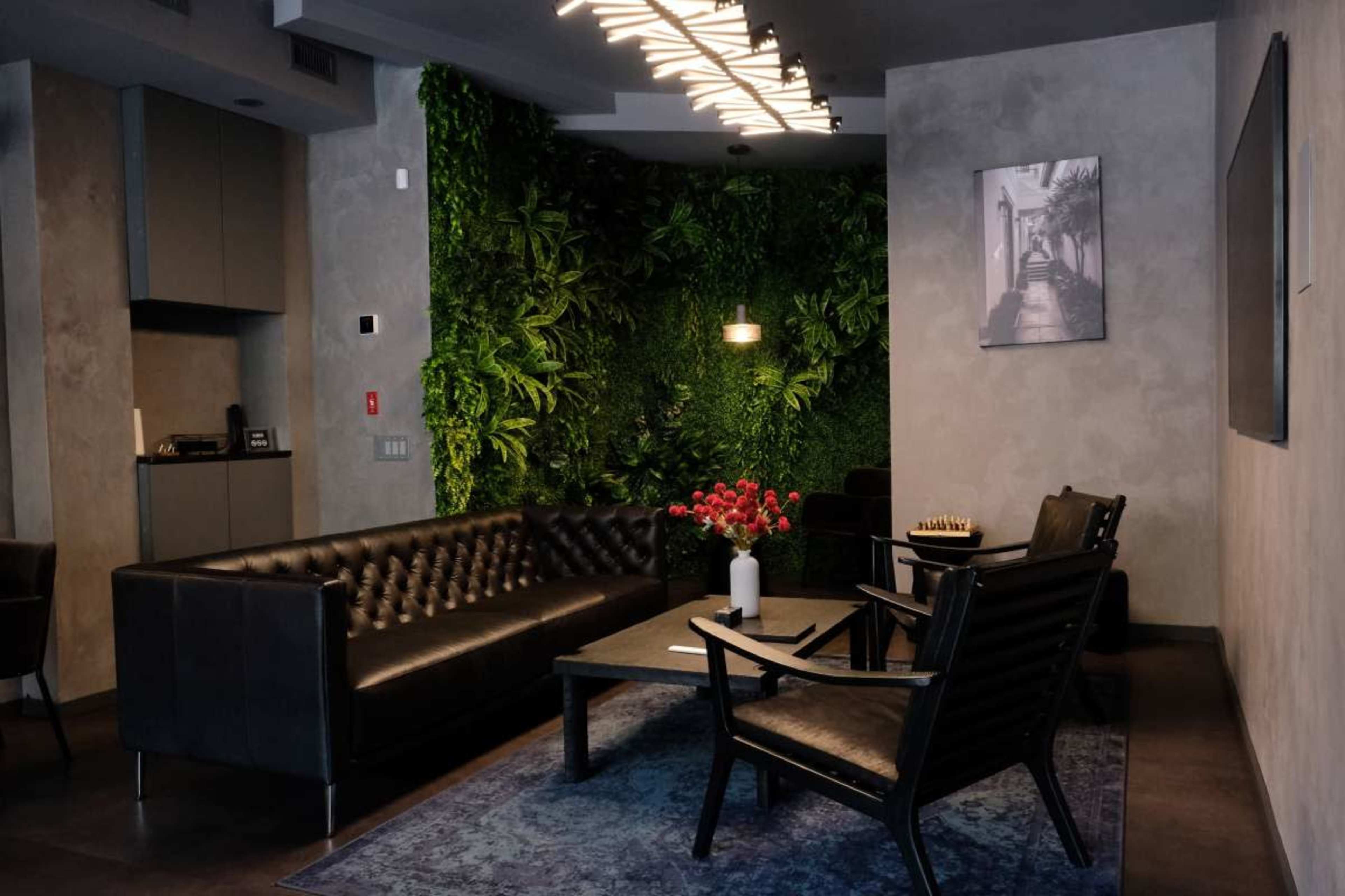 A modern lounge area features a black leather sofa and chairs, a coffee table, and a wall adorned with greenery and a framed photograph.