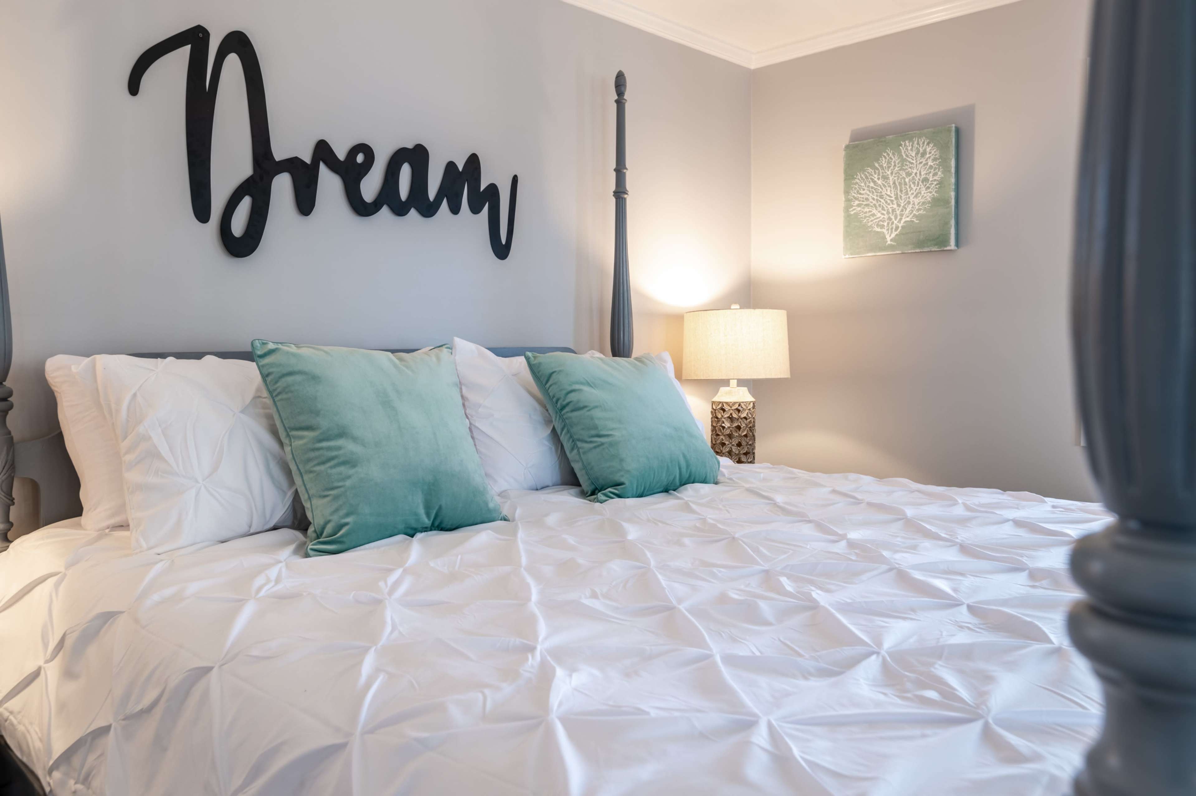 A neatly made bed with a white quilt and decorative pillows, against a gray wall featuring a wooden "Dream" sign and a framed picture of a leaf.