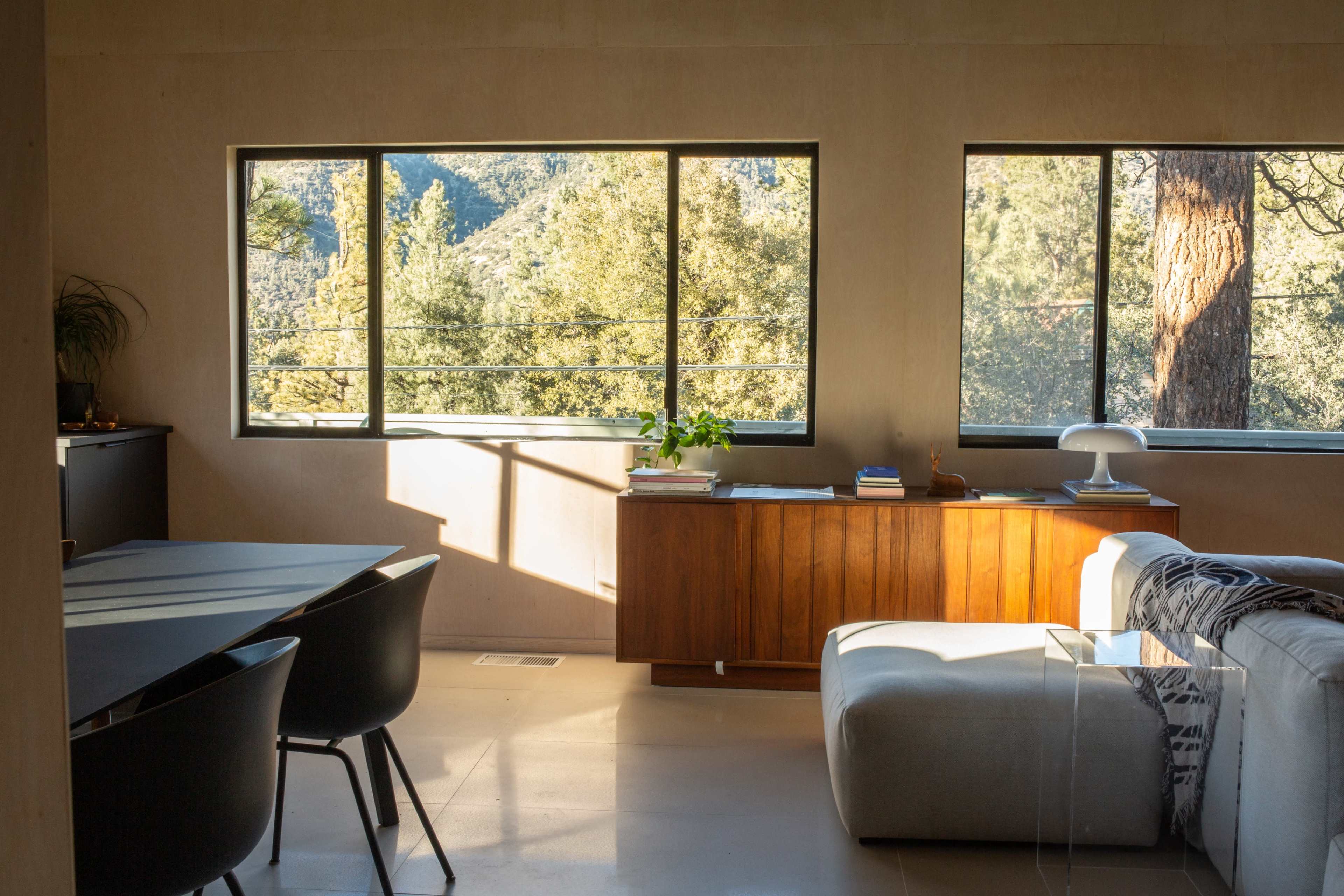 A modern living space features large windows showcasing a natural landscape, with a wooden sideboard and a sectional sofa arranged in the room.