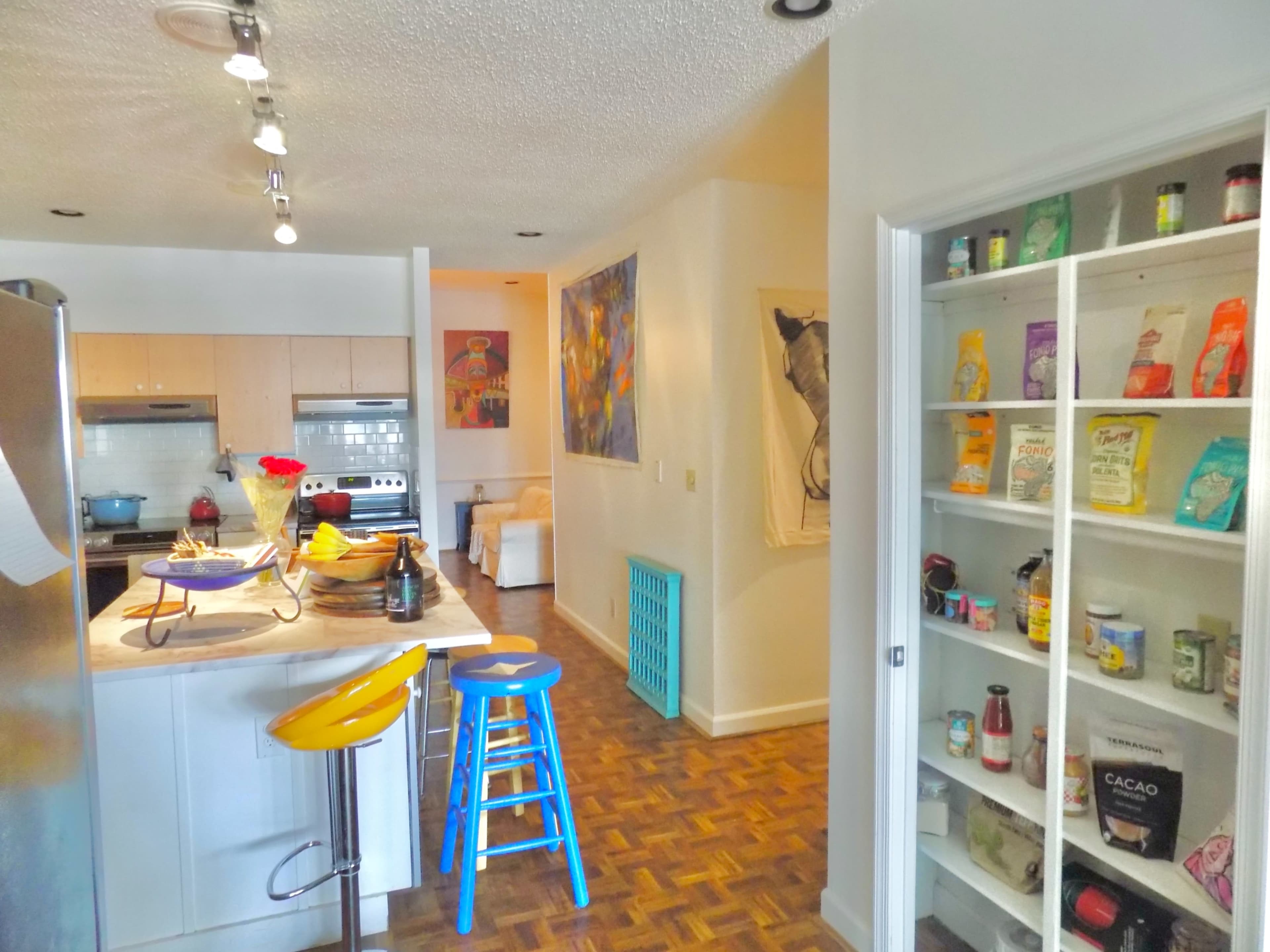 The image shows a kitchen adjacent to a pantry with shelves displaying various food items and a small dining area with a wooden countertop and stools.