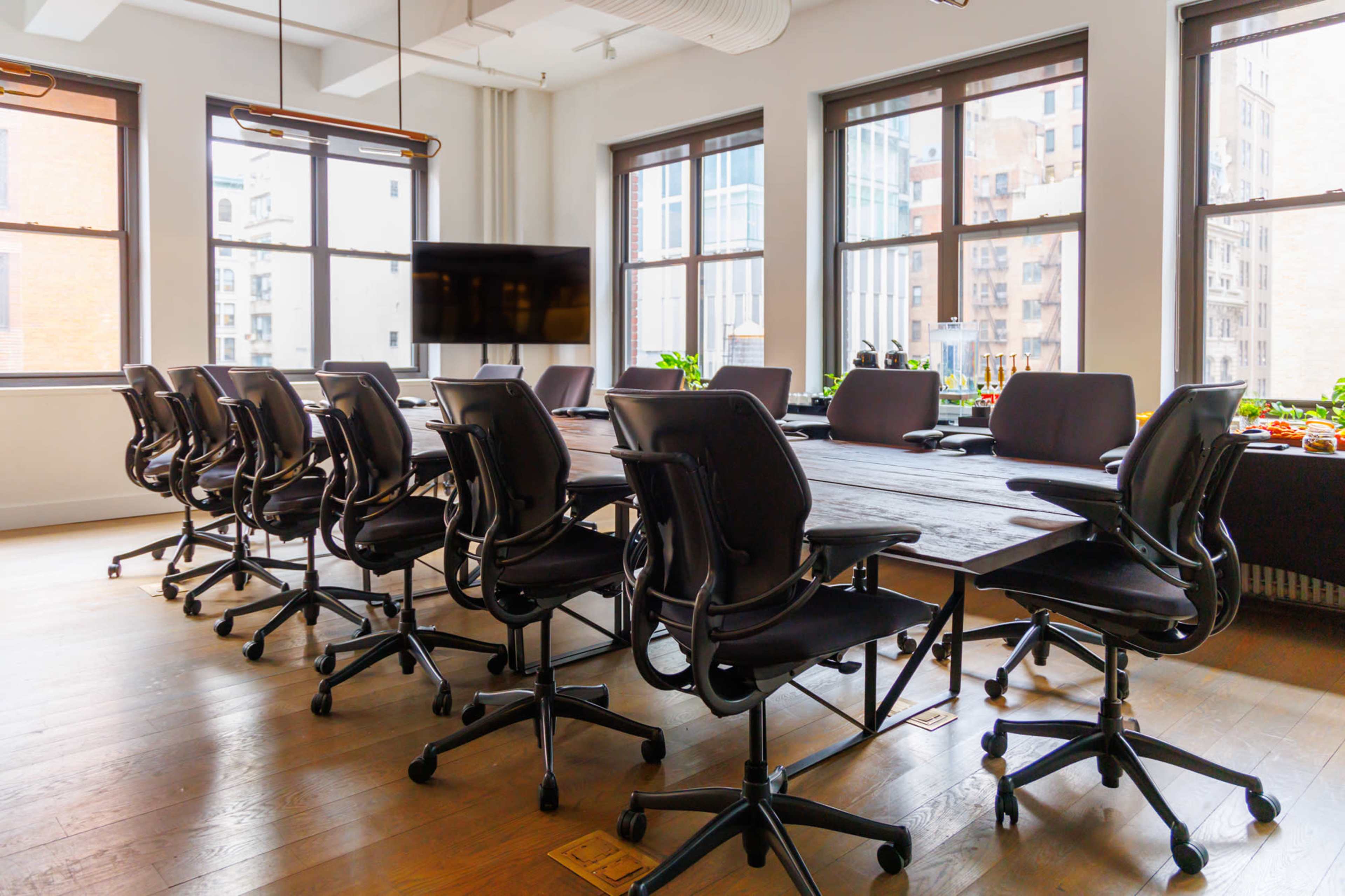 The image shows a modern conference room with a long table, ergonomic chairs, and large windows allowing natural light to enter.