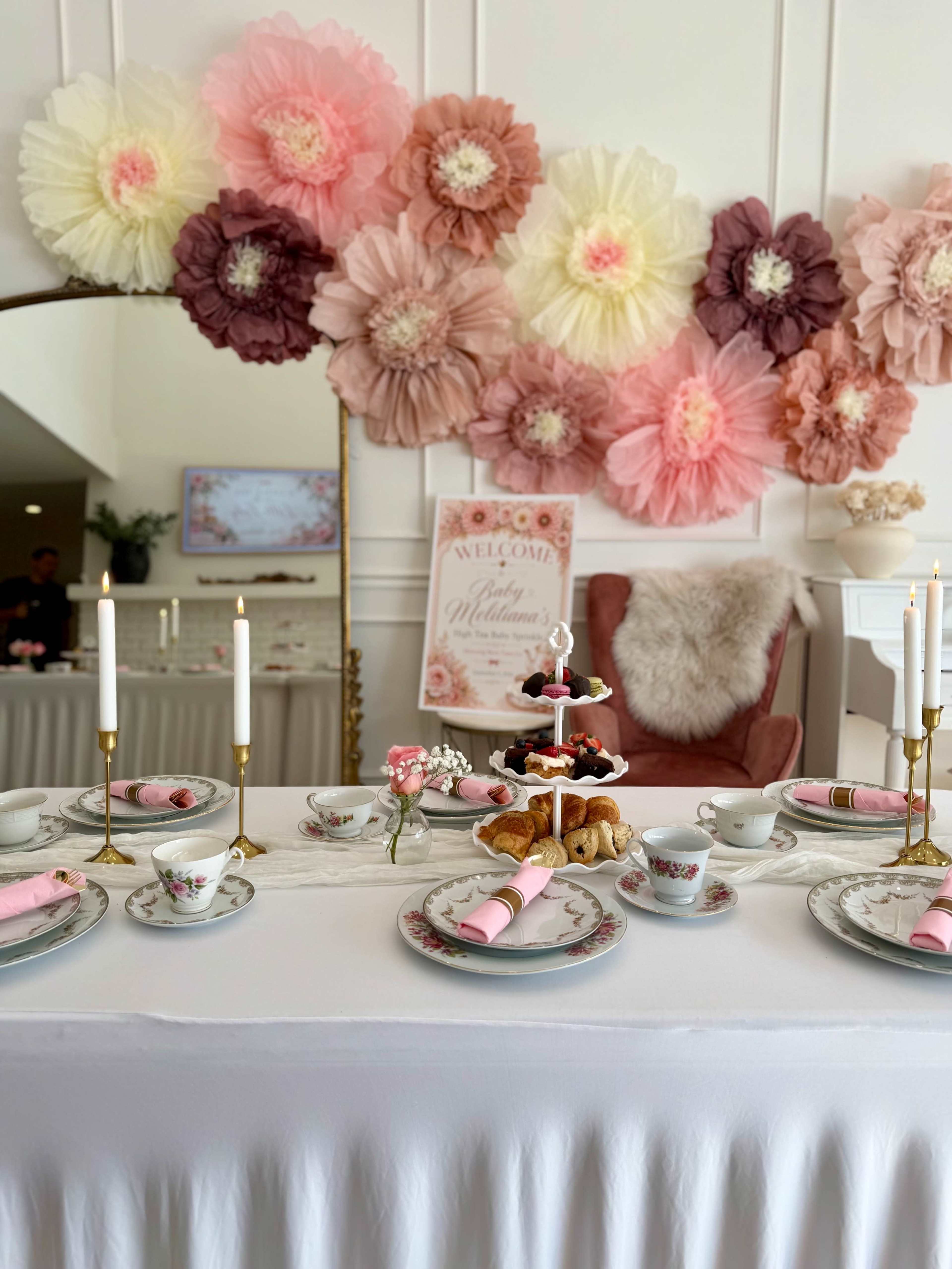 The image features a beautifully arranged tea party table with delicate floral decorations, elegant tableware, and a tiered stand filled with pastries, set against a backdrop of large, colorful paper flowers.
