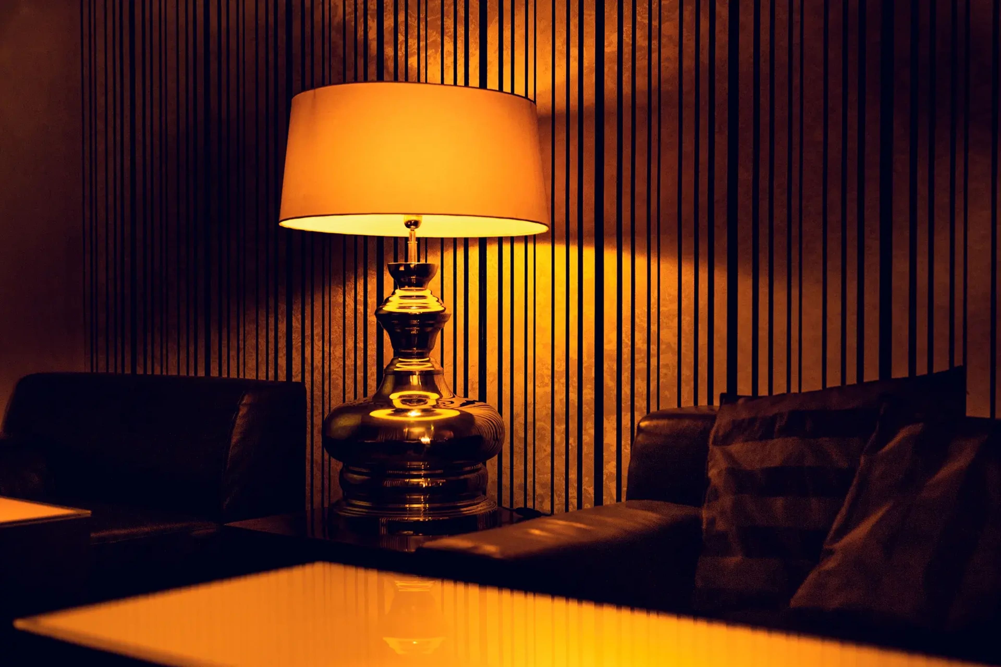 A vintage lamp with a beige shade casts warm light over a black leather sofa in a dimly lit room with striped walls.