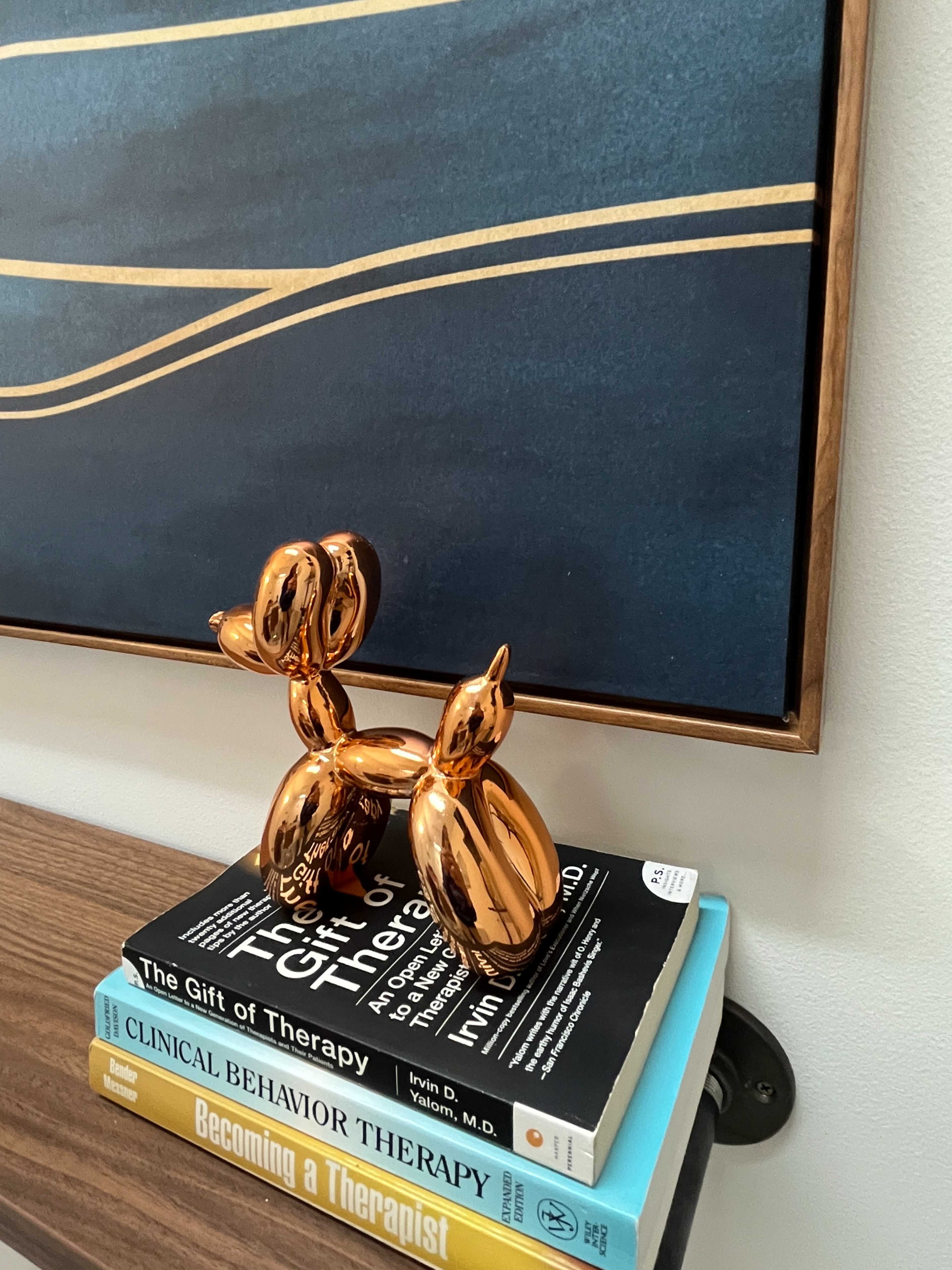 A shiny, balloon-like dog sculpture sits atop a stack of books on a shelf, with an abstract framed piece of art in the background.