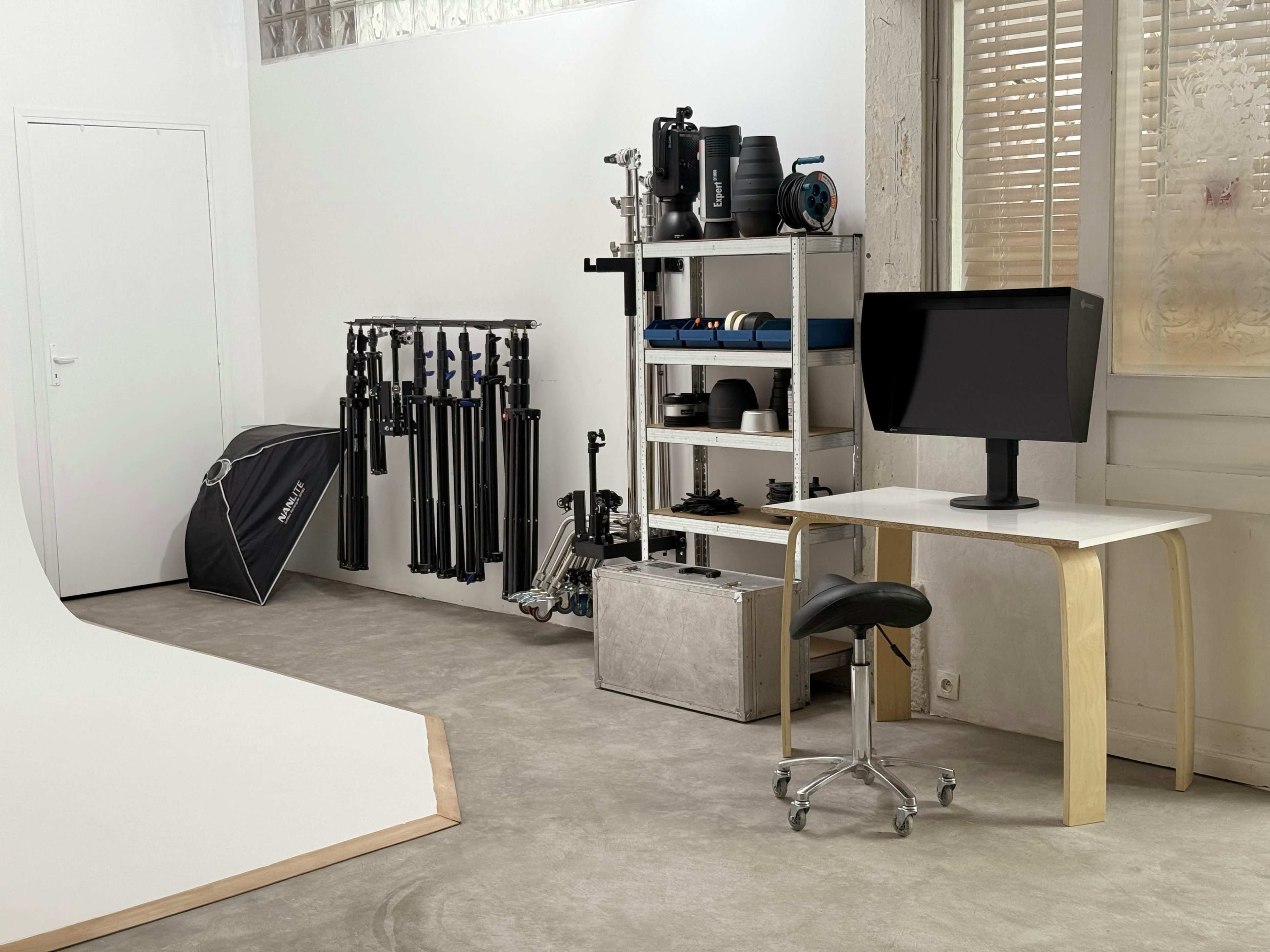 The image showcases a well-organized photography studio featuring a blank backdrop, a monitor on a desk, and various photography equipment stored on shelves.