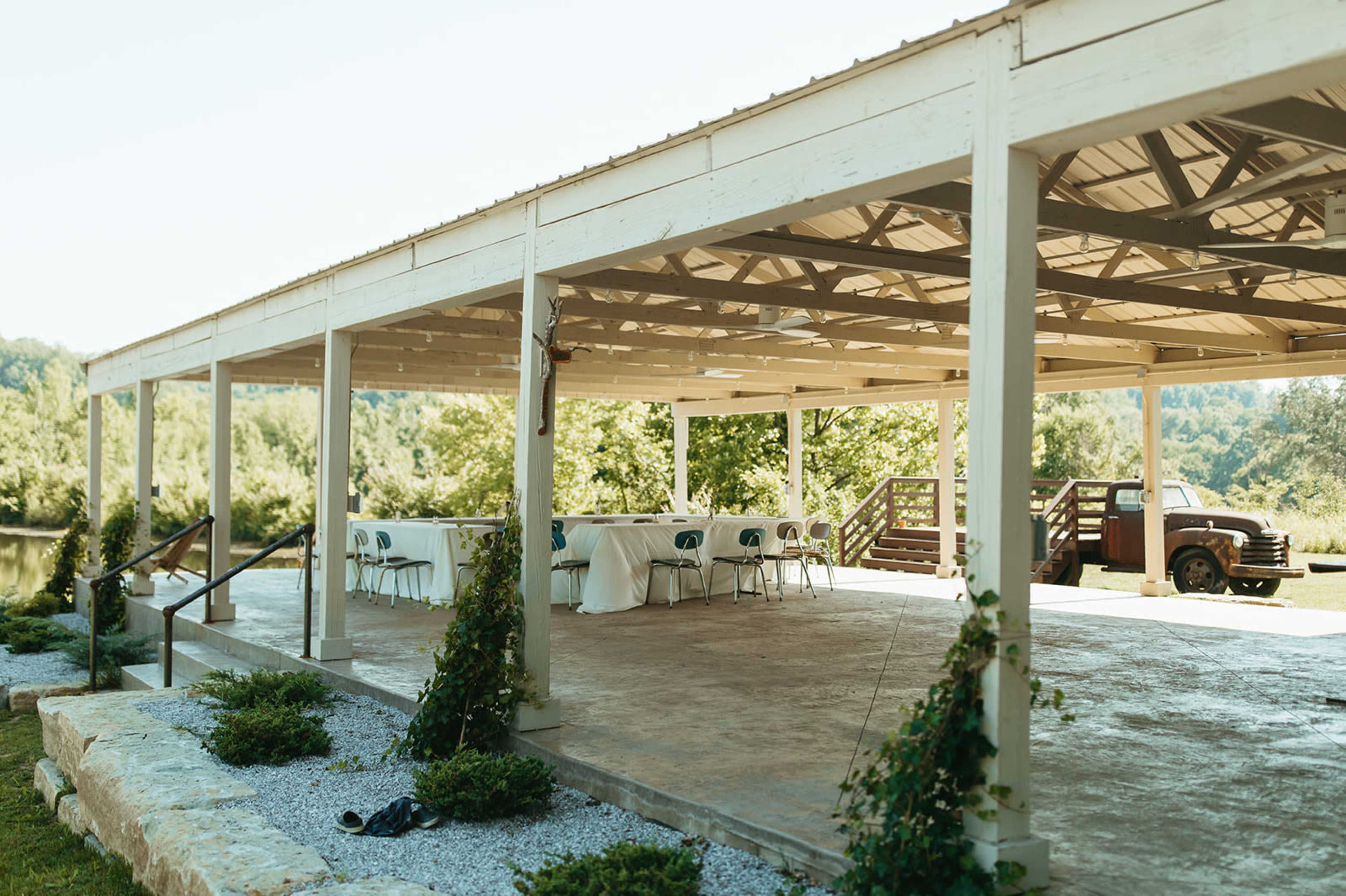 A covered pavilion with tables and chairs set up is situated next to a grassy area and a pond, with a vintage truck parked nearby.
