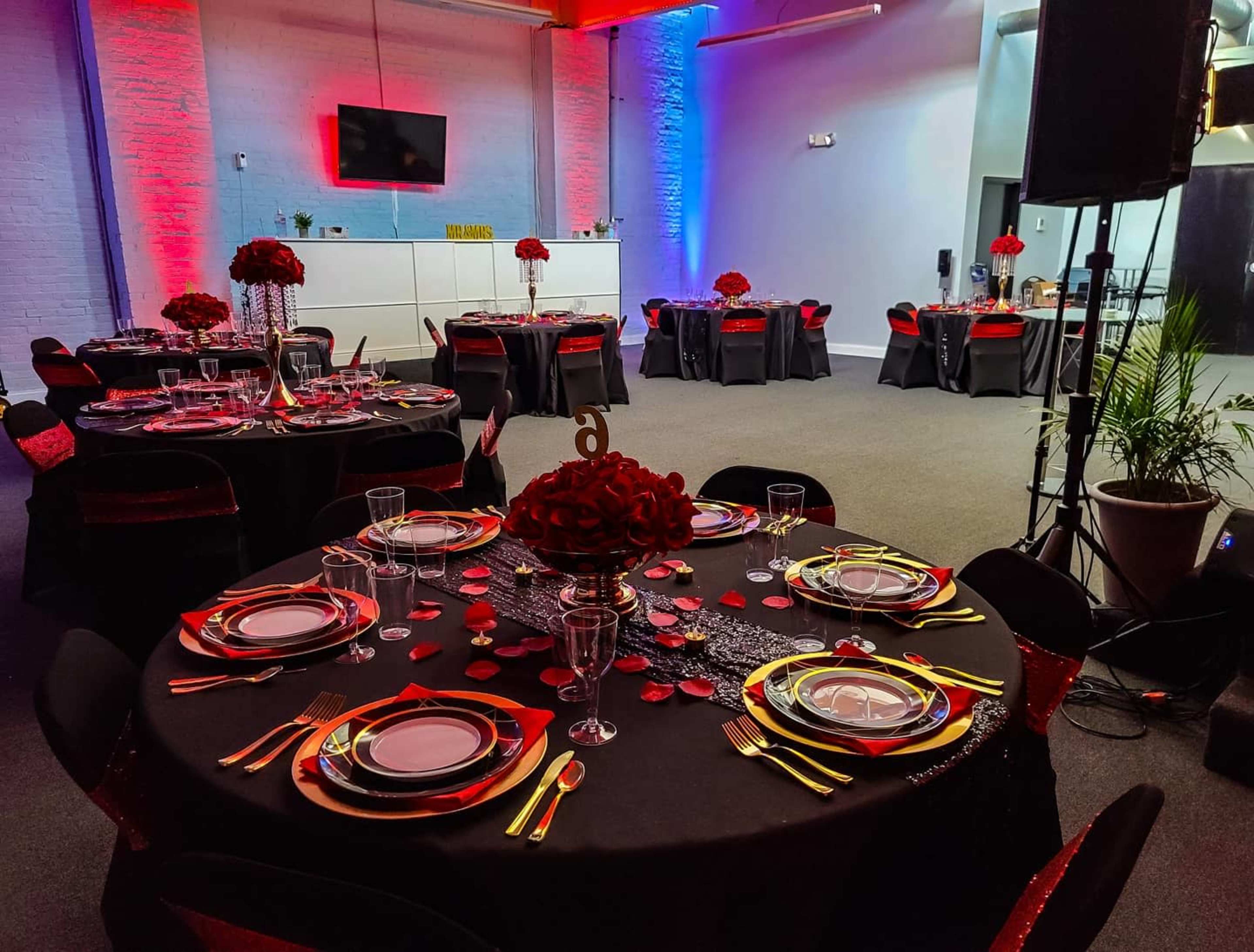 A spacious event venue is set up with round tables covered in black cloth, adorned with red floral centerpieces and elegant table settings, under vibrant red and blue lighting.