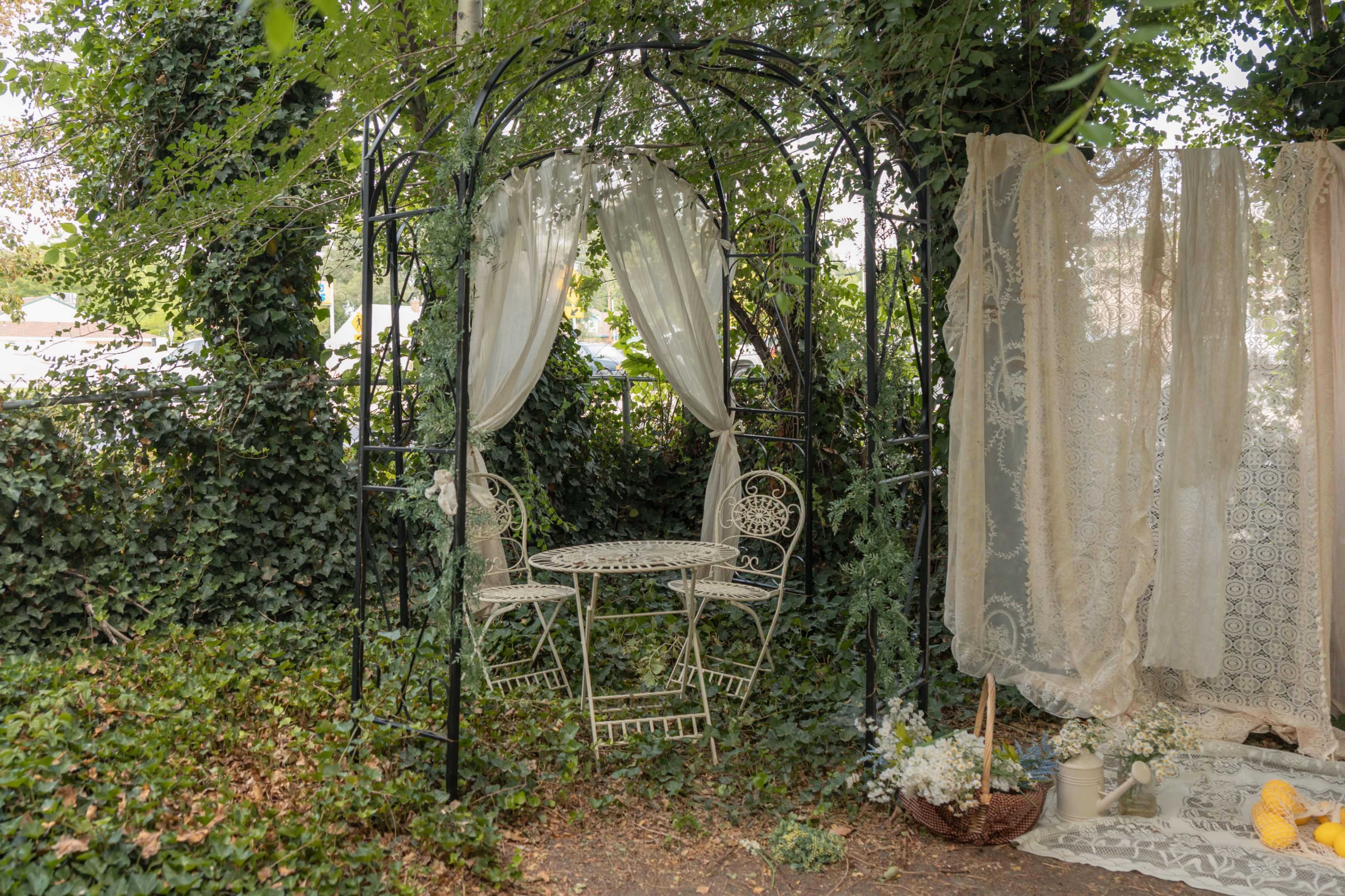A wrought iron gazebo with a small table and chairs is partially covered by sheer curtains and surrounded by greenery and flowers.