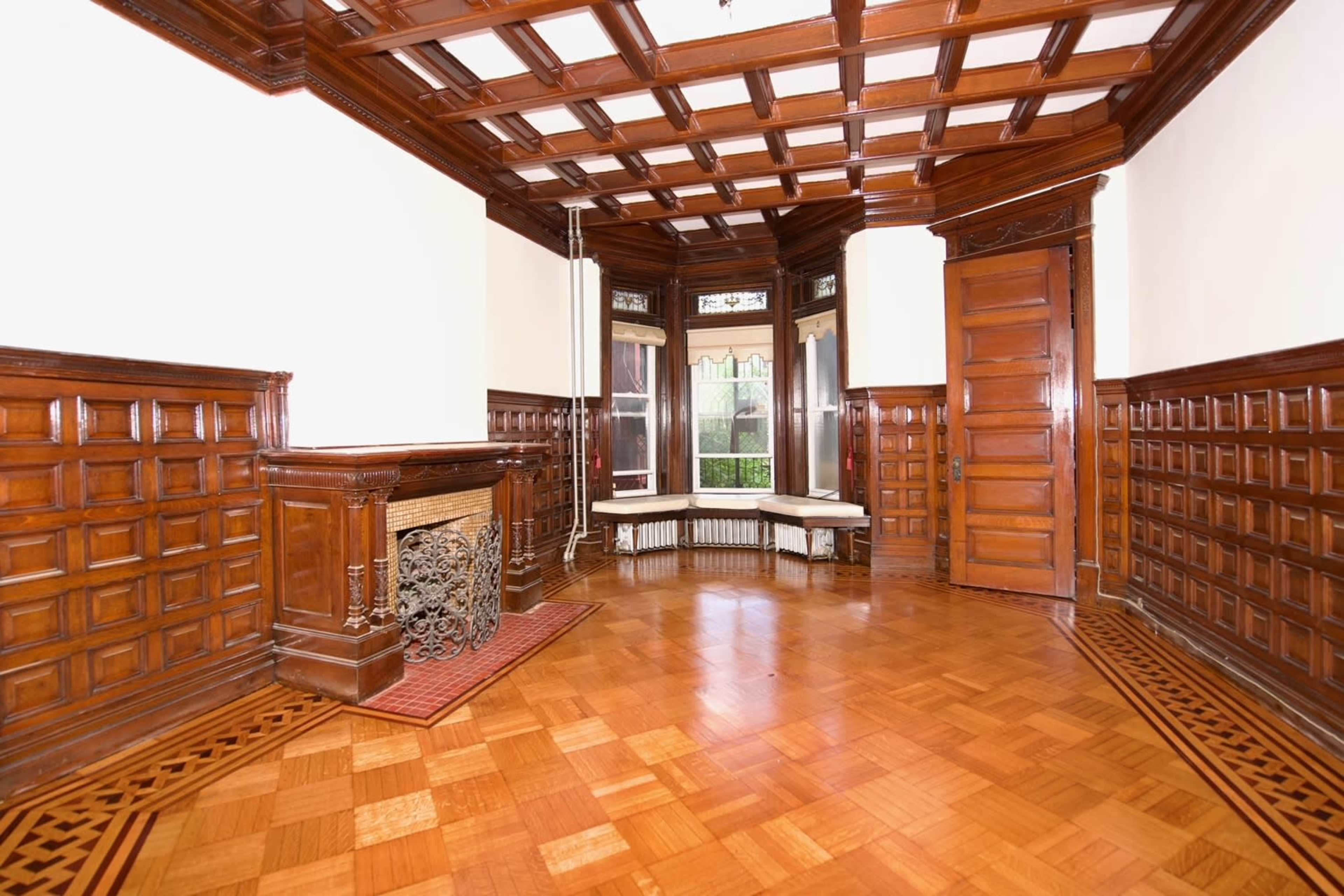 A wooden-paneled room with a coffered ceiling, featuring a fireplace and three large windows.