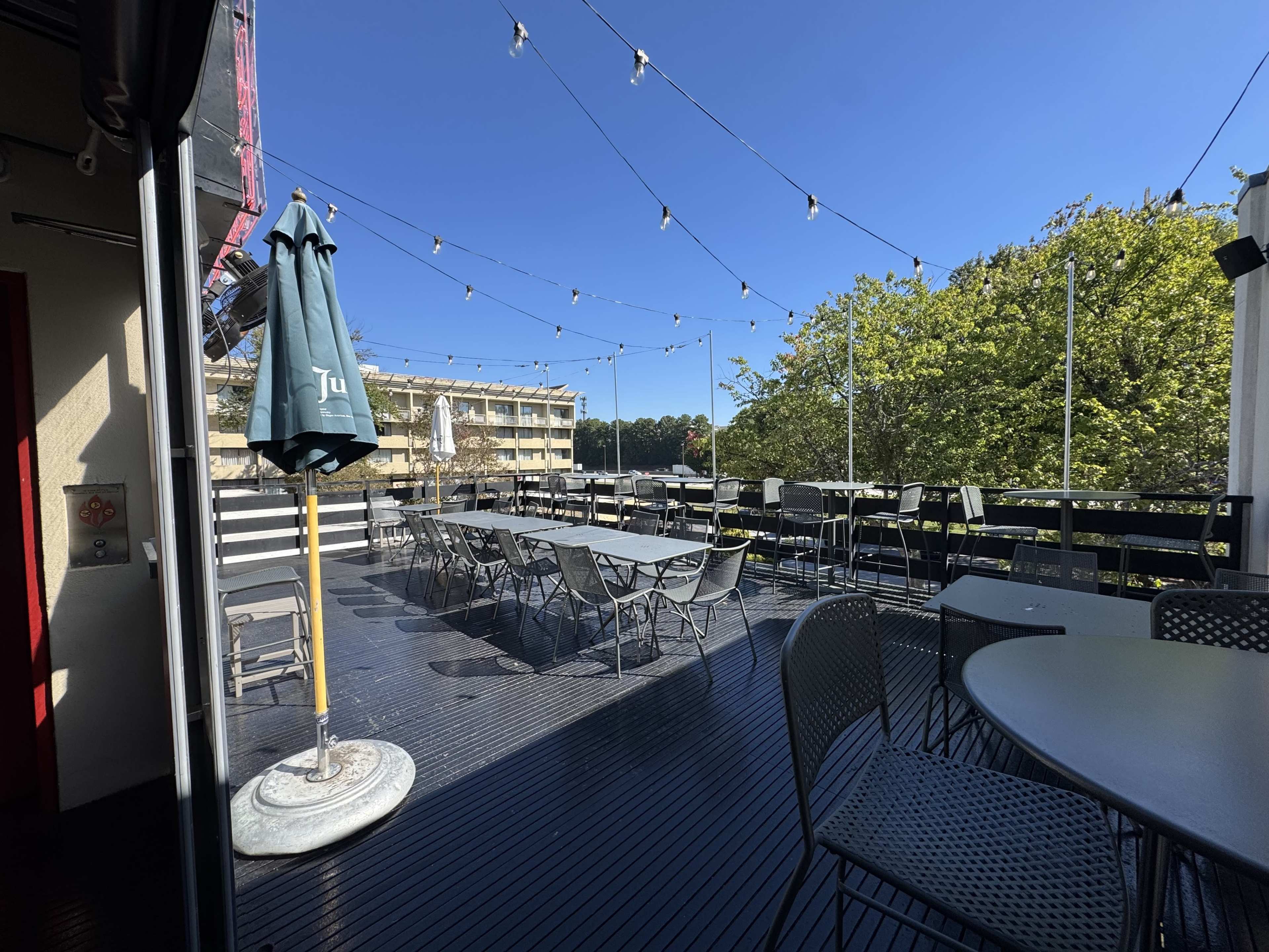 The image shows an outdoor patio area with tables and chairs, string lights overhead, and greenery visible in the background under a clear blue sky.