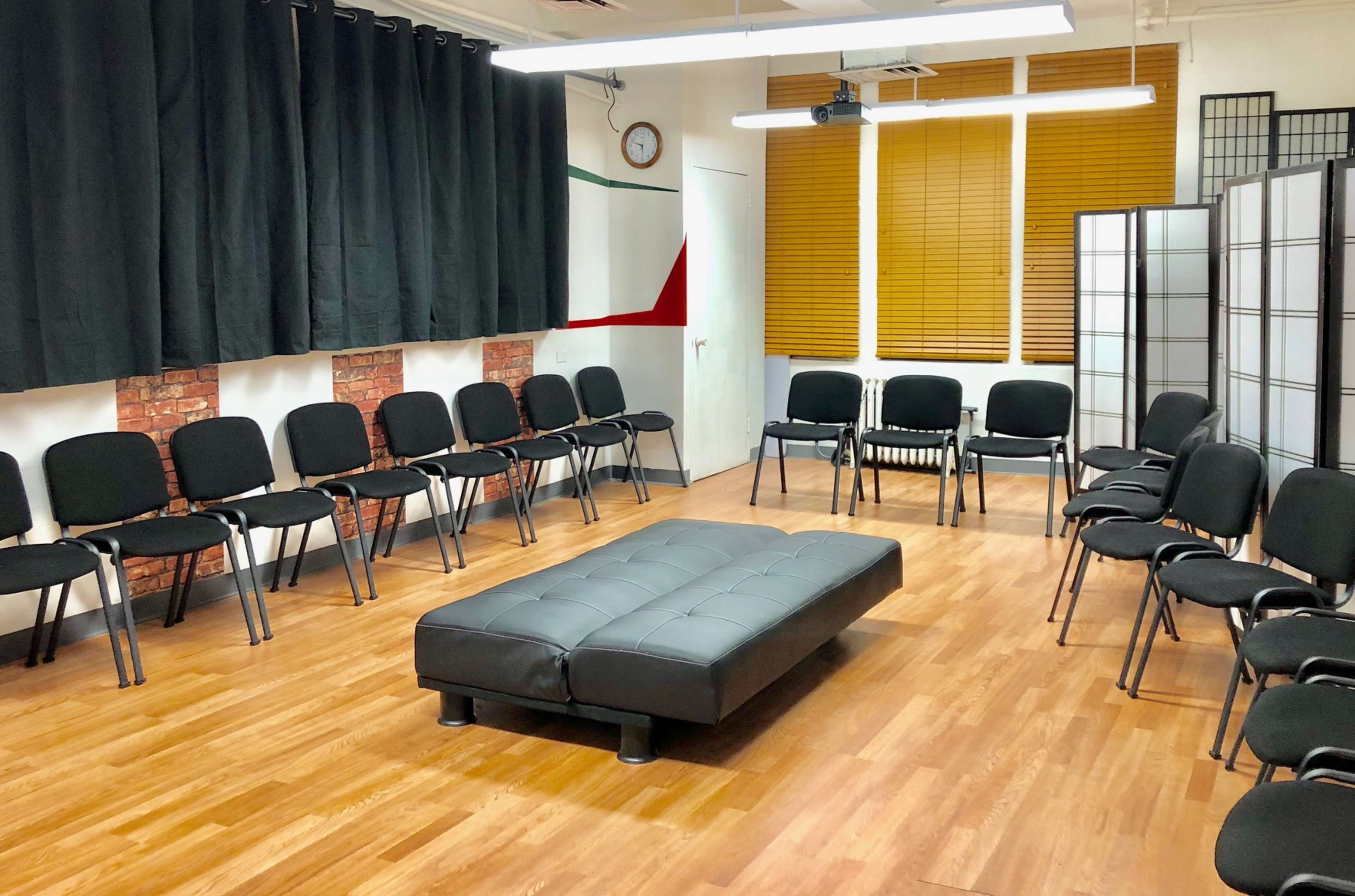 A room arranged with chairs in a circular formation around a rectangular black couch, with wooden flooring and partially closed blinds on the windows.