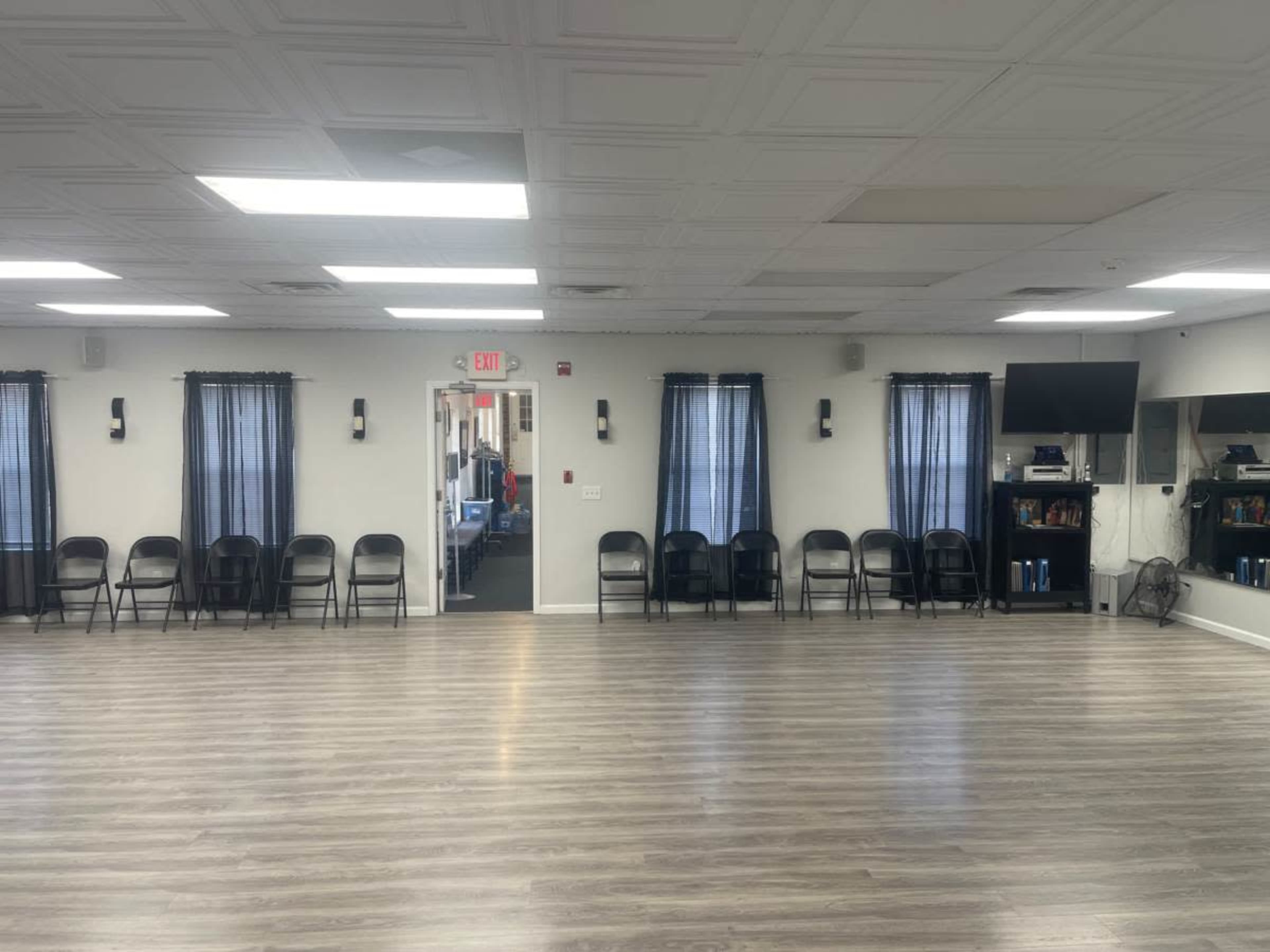 The image shows a spacious room with several black chairs lined up against the walls, large windows covered with dark curtains, and a small television mounted on one wall.