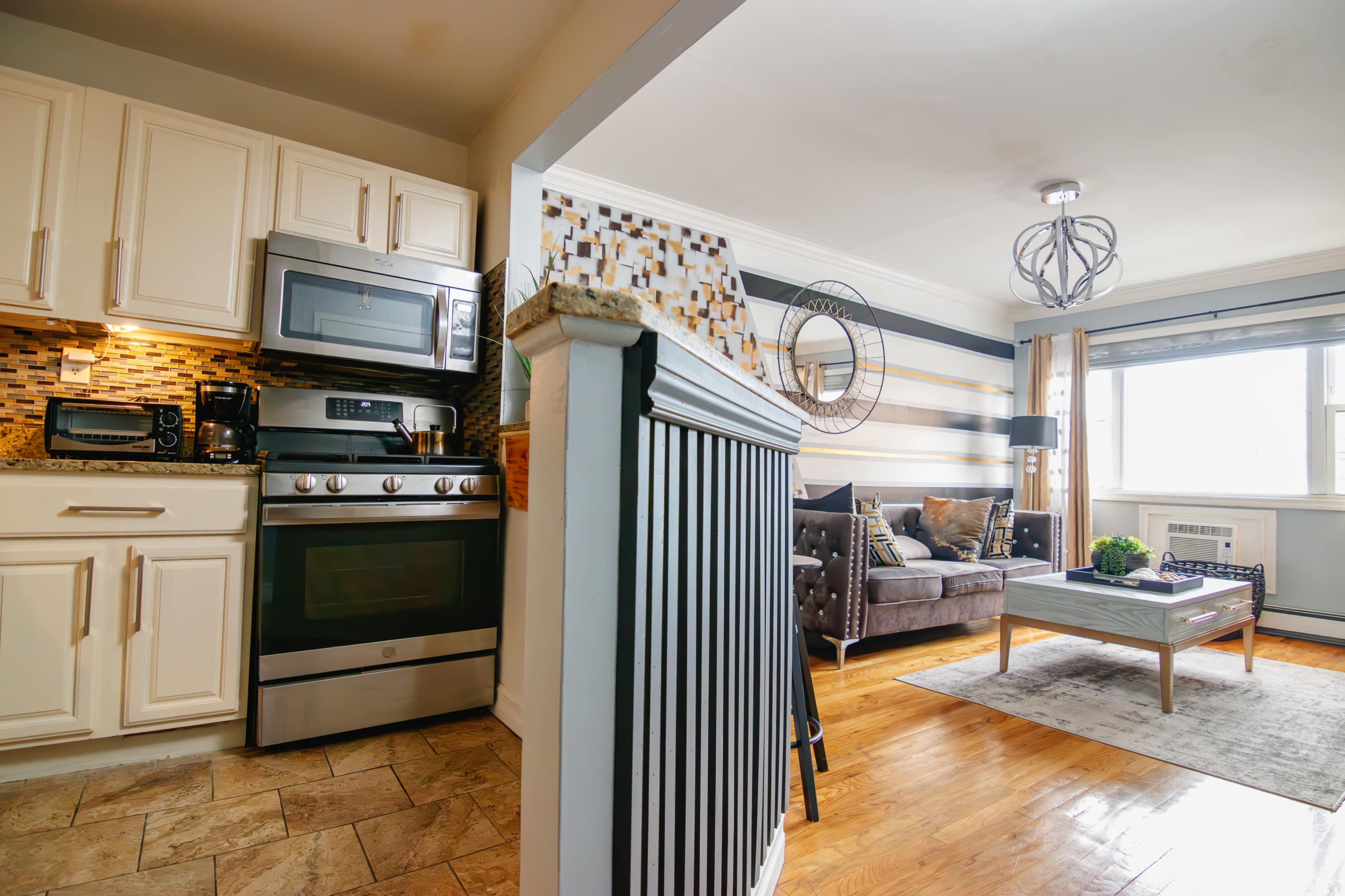 A modern kitchen with light-colored cabinetry and stainless steel appliances that opens up to a living area featuring a gray sofa and a coffee table.