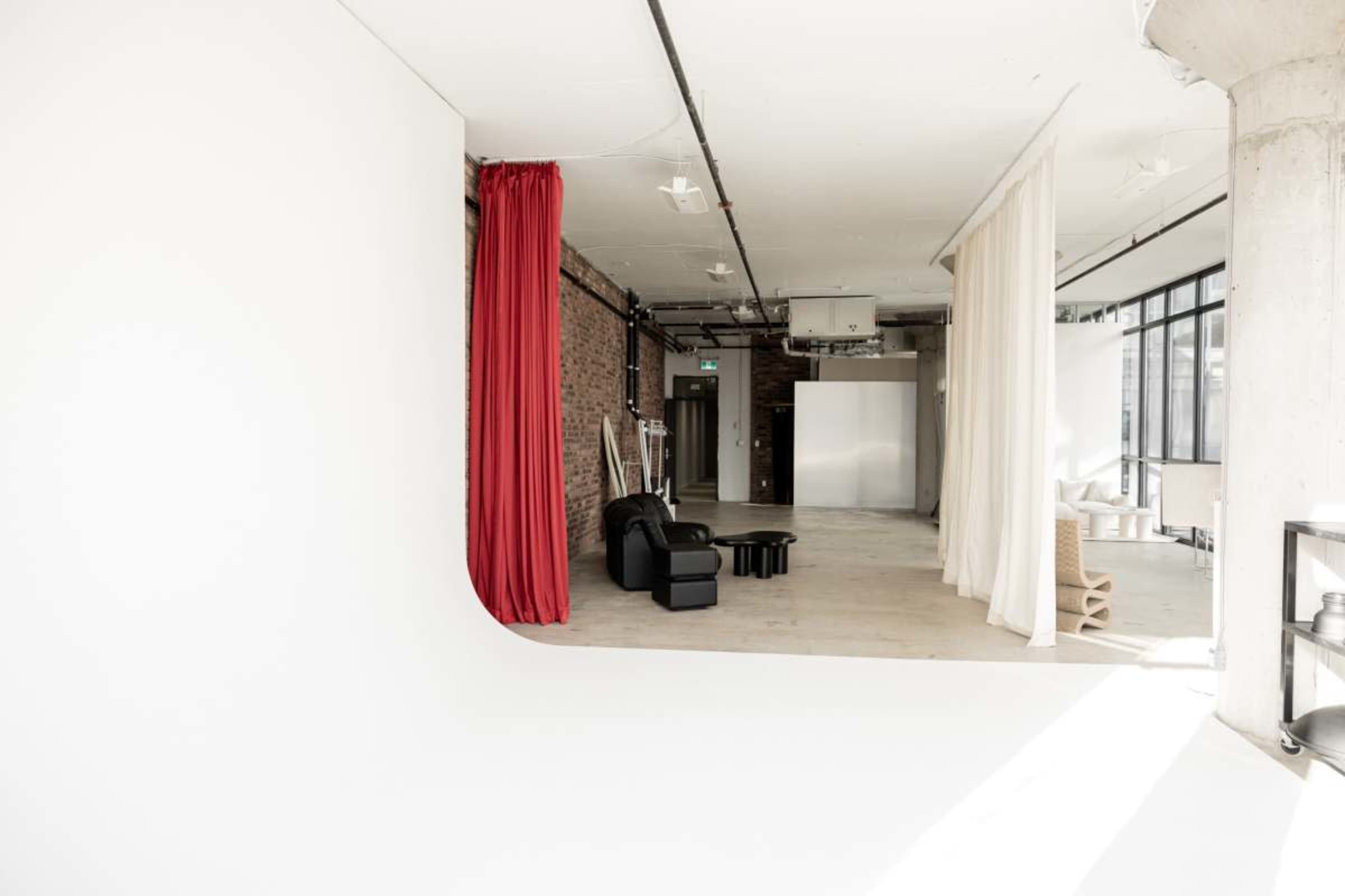 The image shows a spacious, modern studio with white walls, exposed brick, and a black sofa, separated by draped fabric panels.