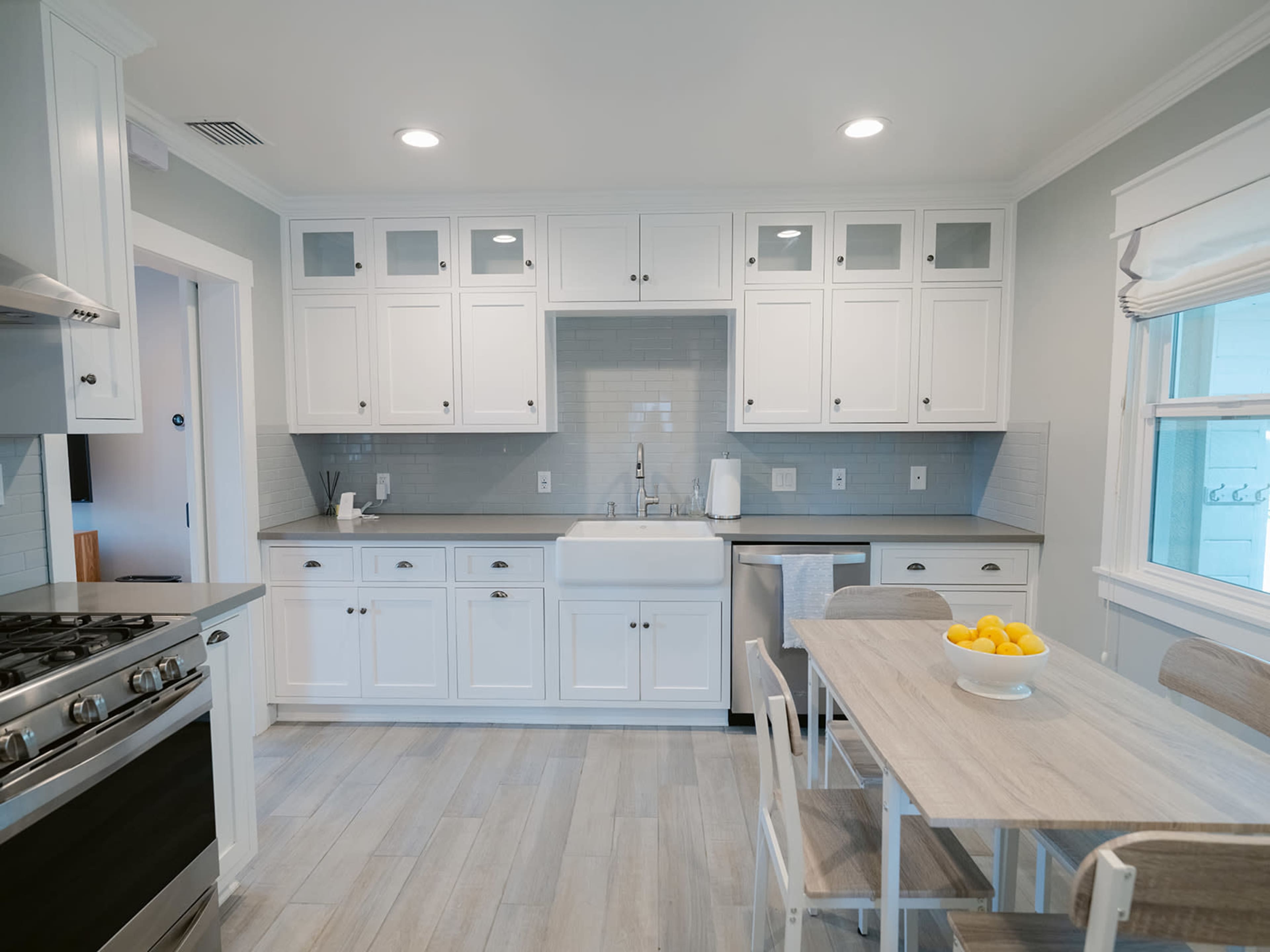 The image shows a modern kitchen with white cabinetry, a farmhouse sink, and a dining table with a bowl of lemons.
