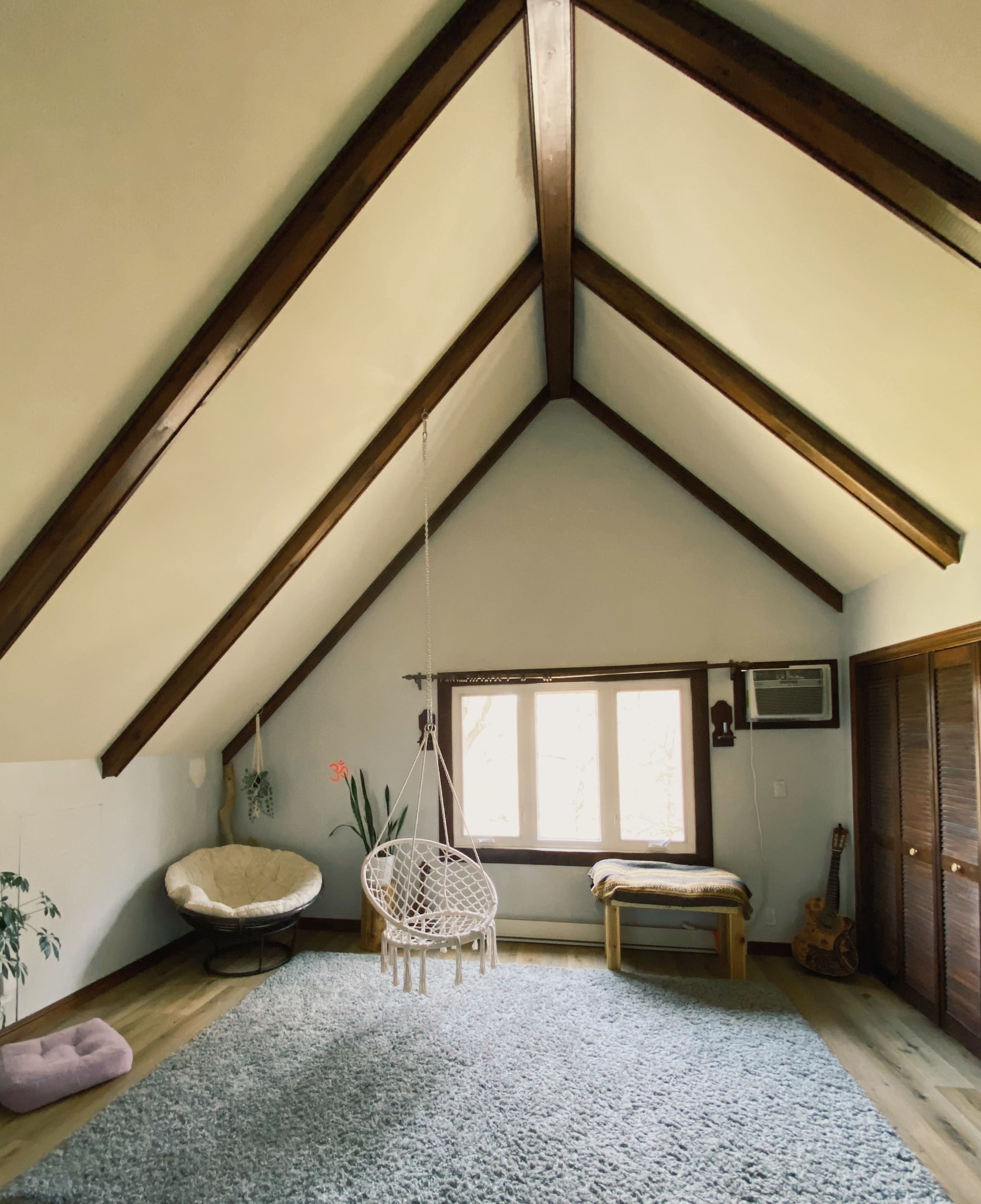 The room features a high, sloped ceiling with wooden beams, a hanging chair, and a large window allowing natural light to enter.