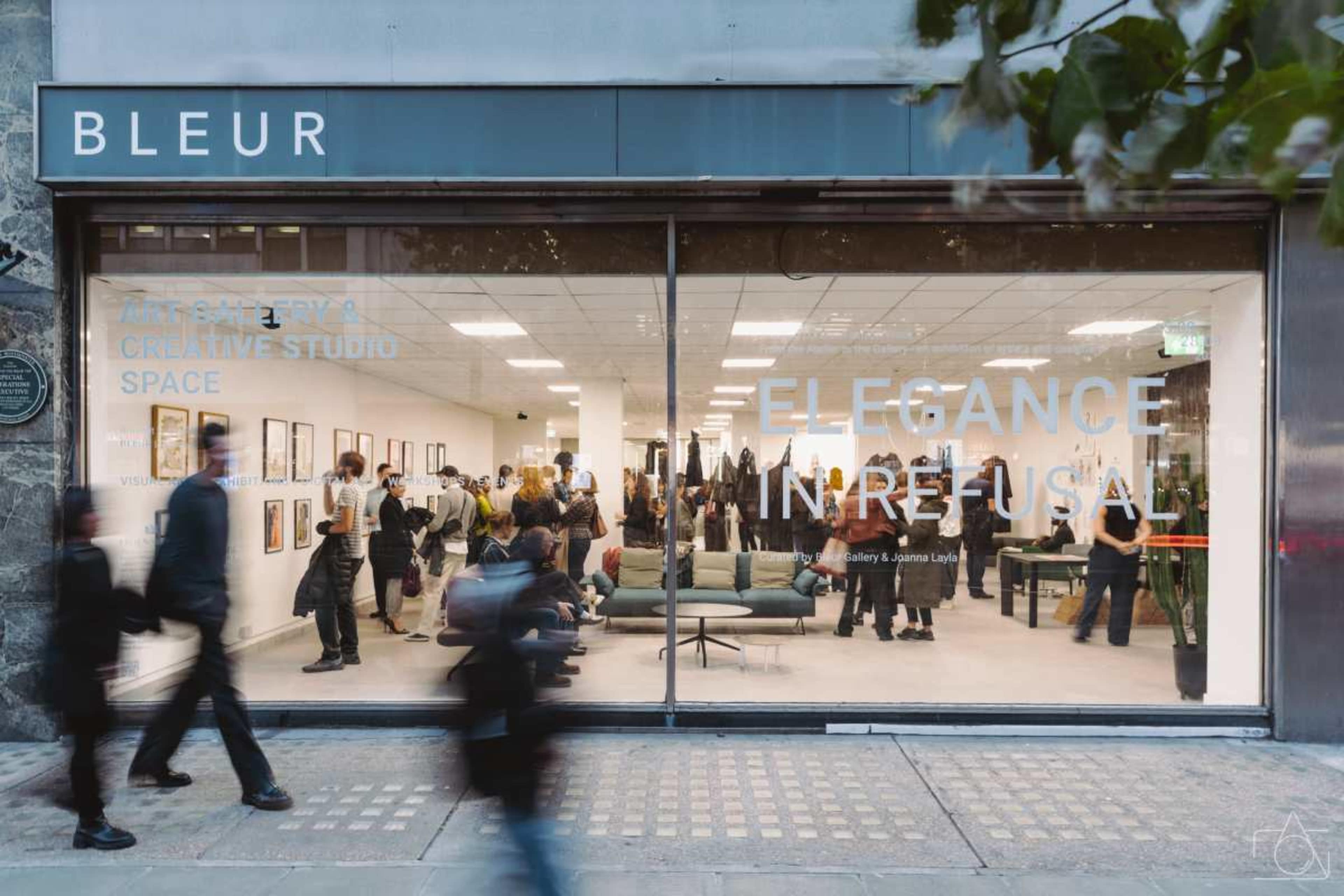 The image shows a modern art gallery with large windows displaying the words "Bleur" and "Elegance in Reusal," where a crowd is gathered inside and outside, viewing artworks and engaging in conversation.