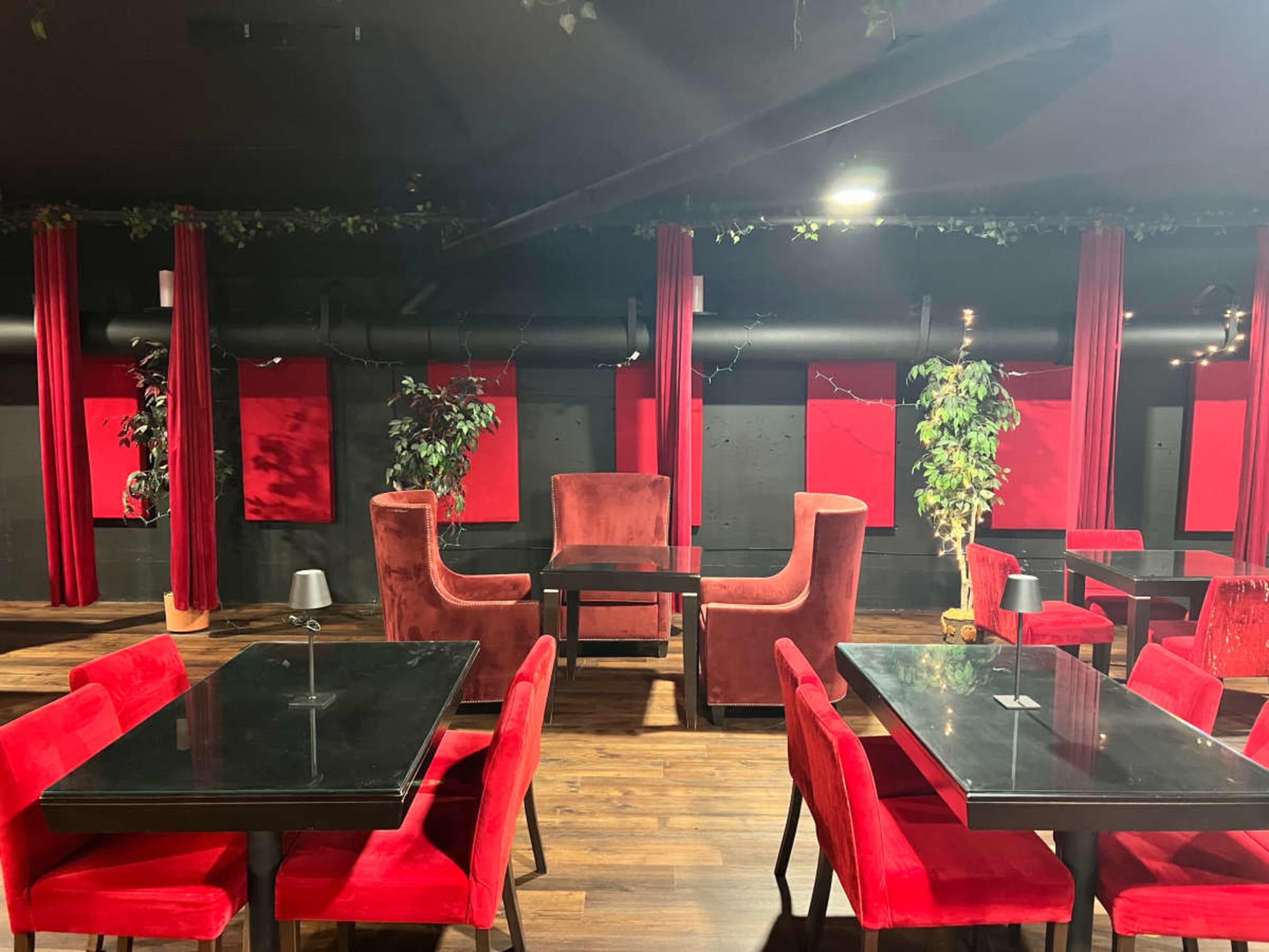 The image shows a dimly lit lounge featuring red velvet furniture, including chairs and booths, paired with black tables and greenery.