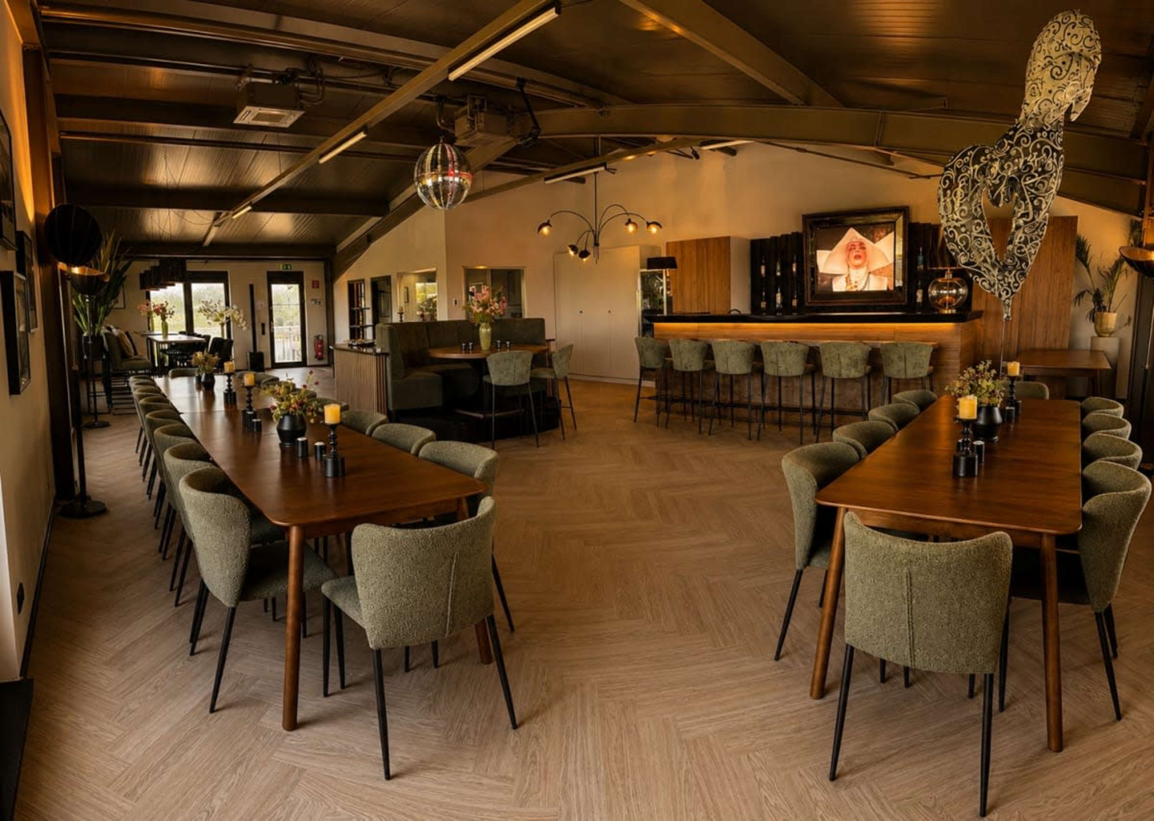 The image shows an interior of a dining space with multiple large tables arranged neatly, surrounded by upholstered chairs and featuring a bar area in the background.