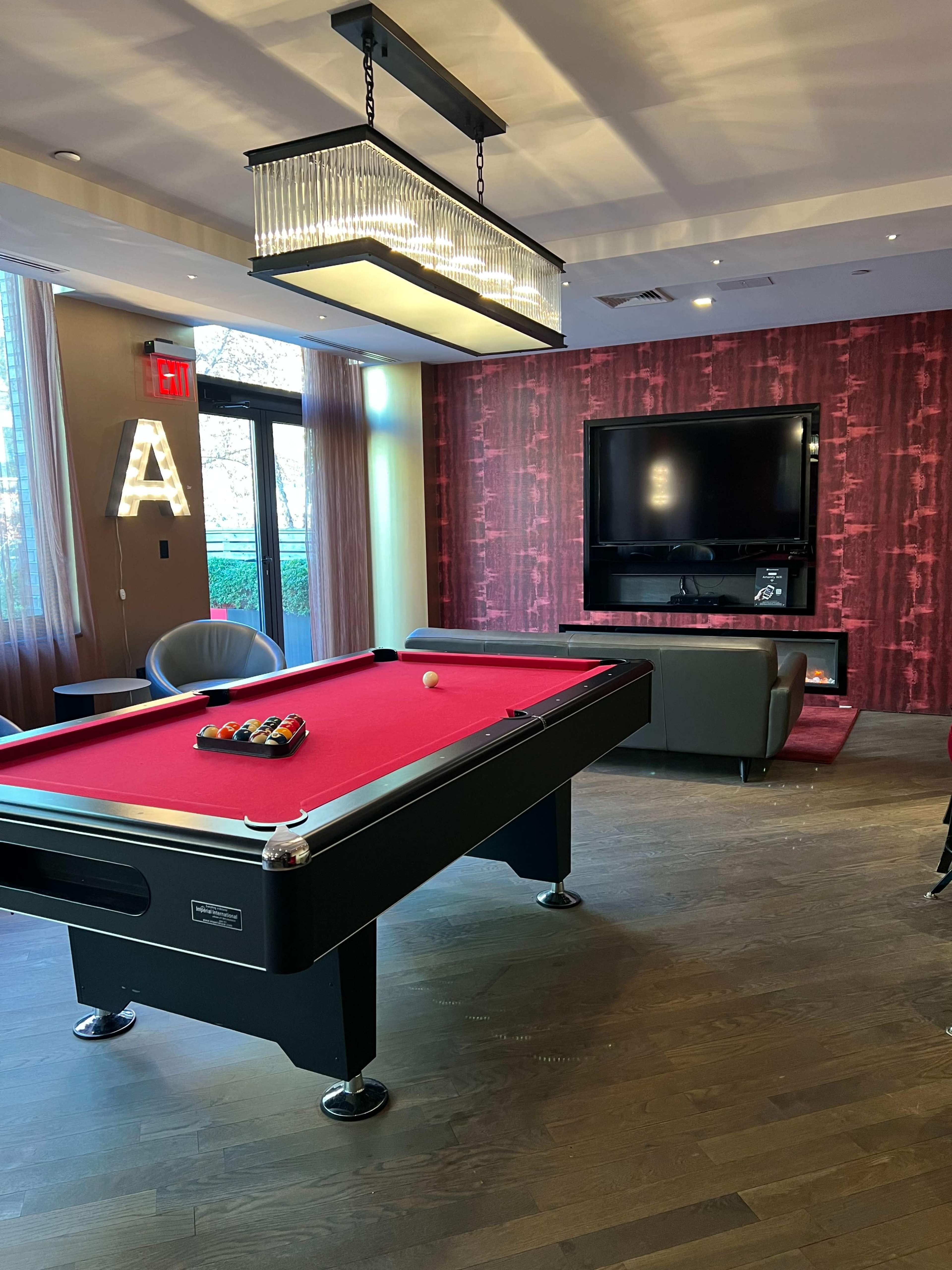 A modern game room featuring a pool table with a red felt surface, a large flat-screen TV on a decorative wall, and a stylish lamp hanging overhead.