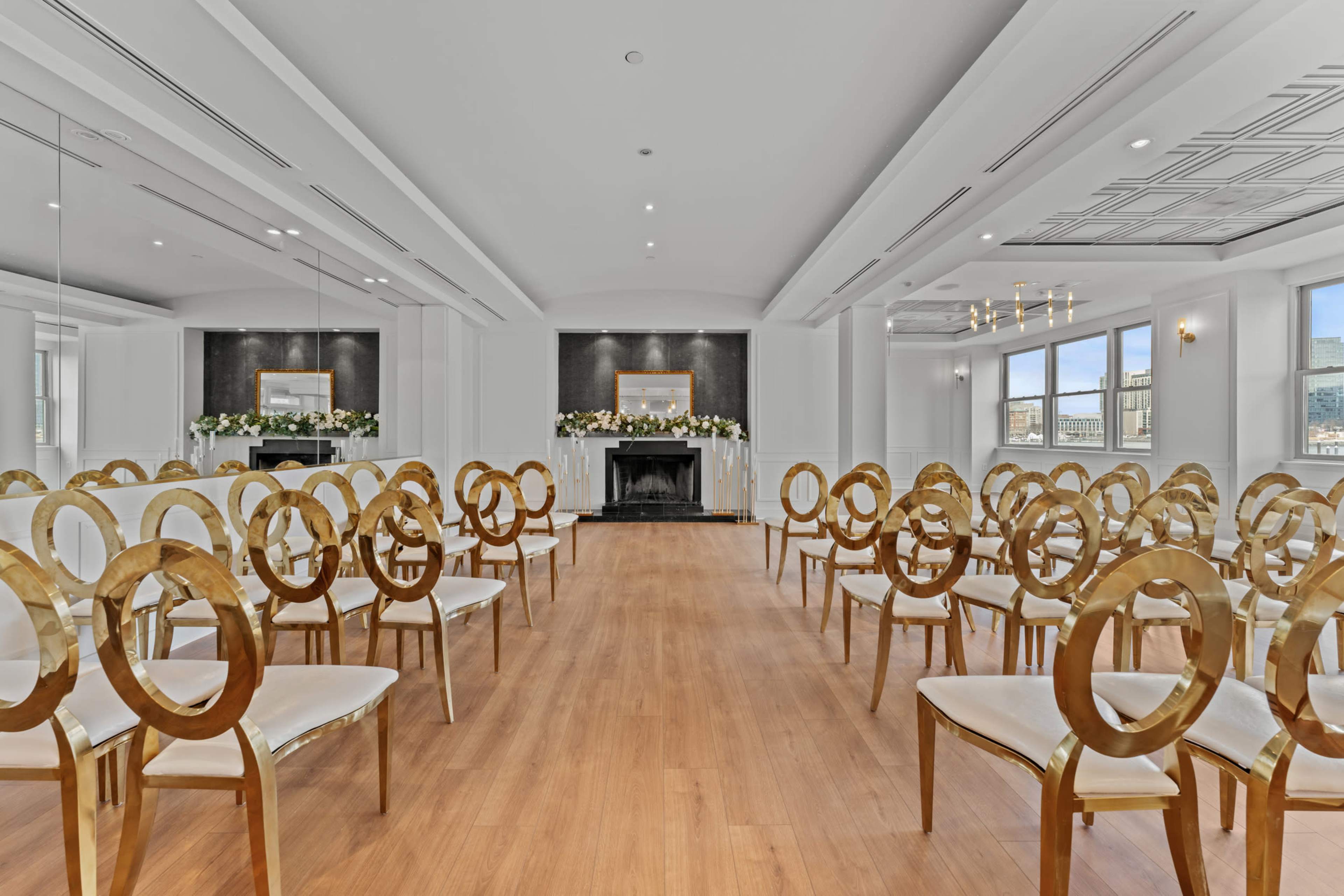 The image shows a spacious, elegant ceremony room arranged with rows of gold-accented chairs facing a fireplace flanked by floral displays.