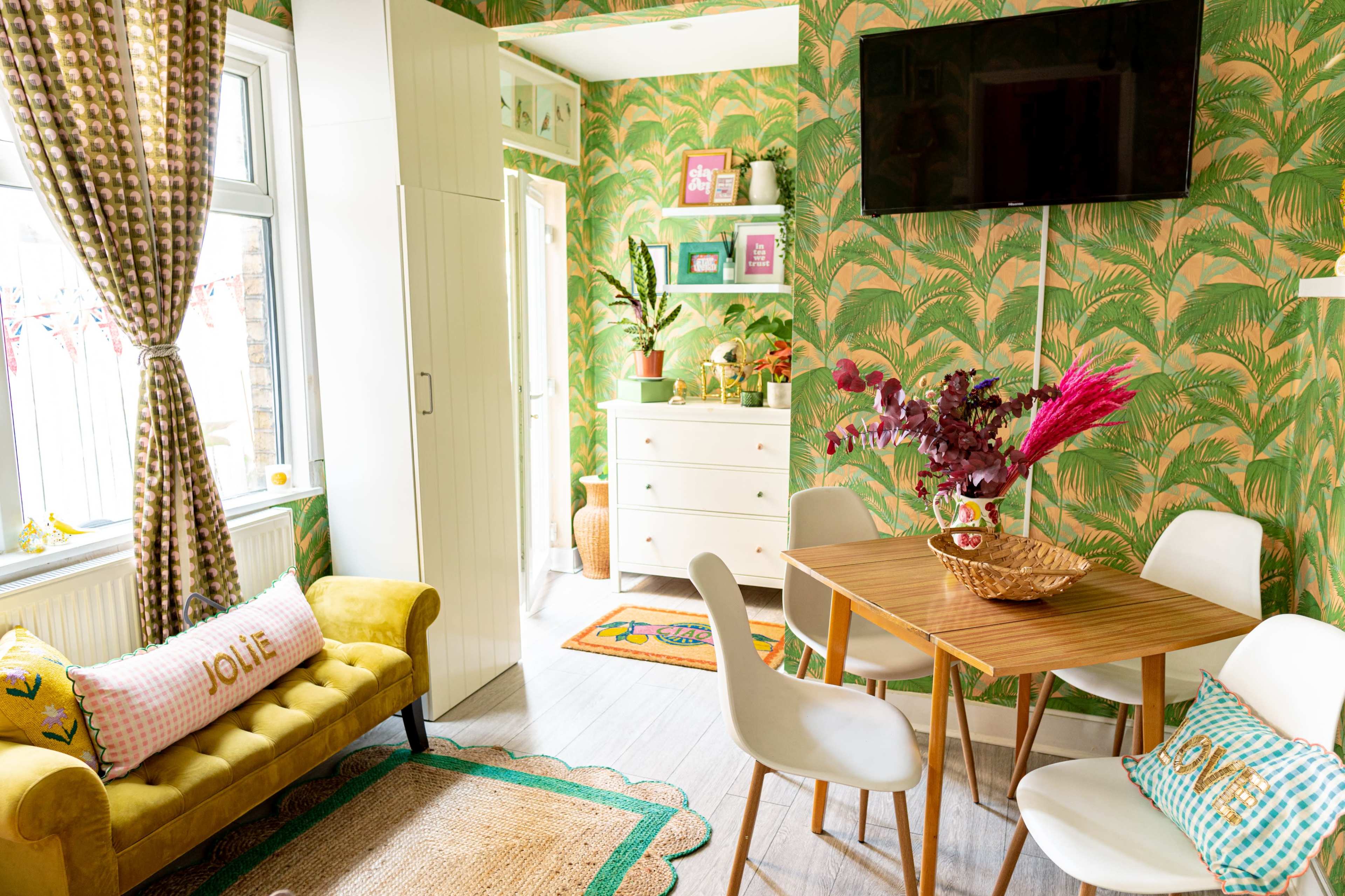 A brightly decorated room features a yellow couch, a wooden dining table with white chairs, and tropical-themed wallpaper.