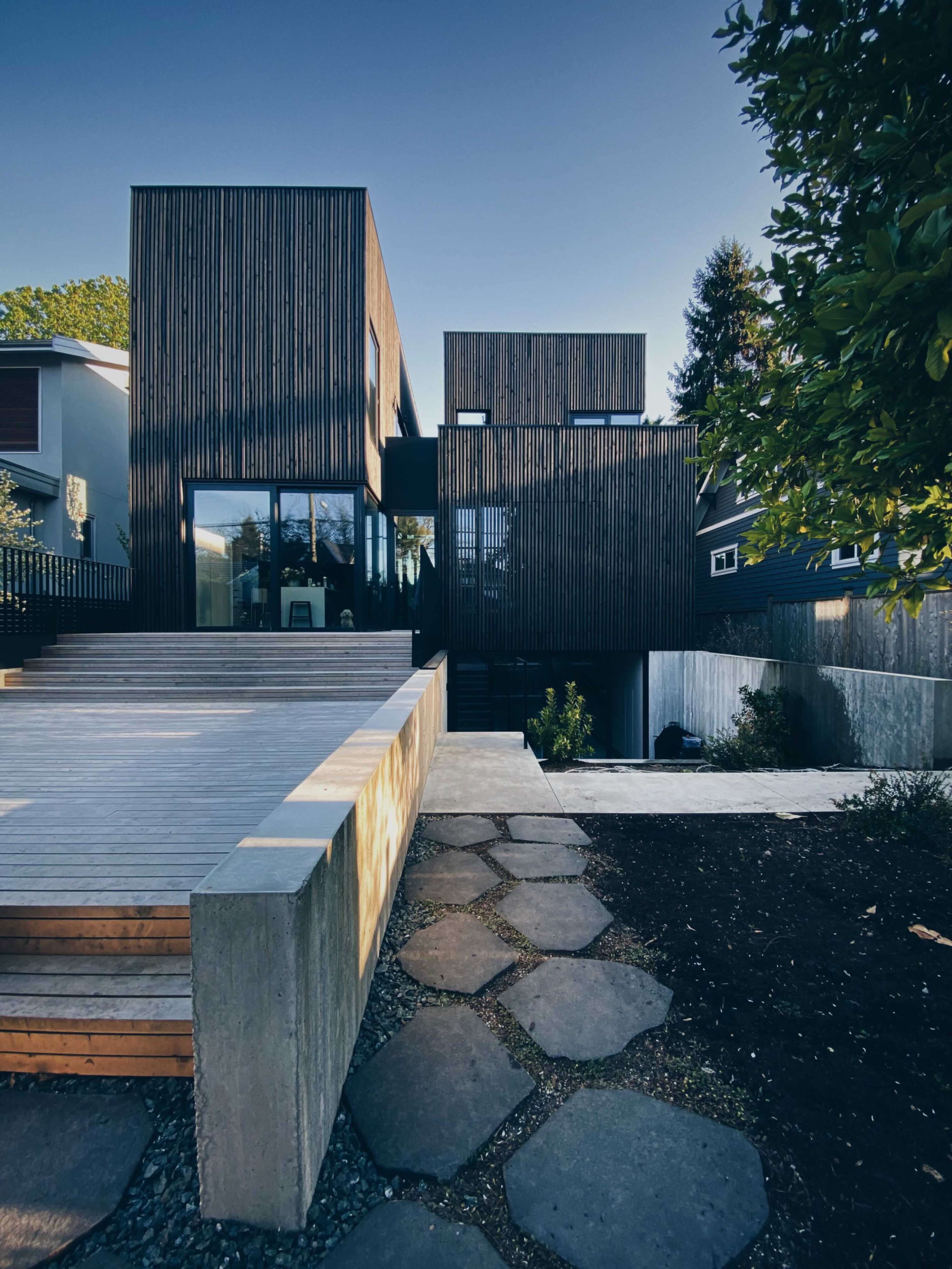 A modern wooden-clad house with large windows, surrounded by a landscaped yard featuring stone pathways and a deck.