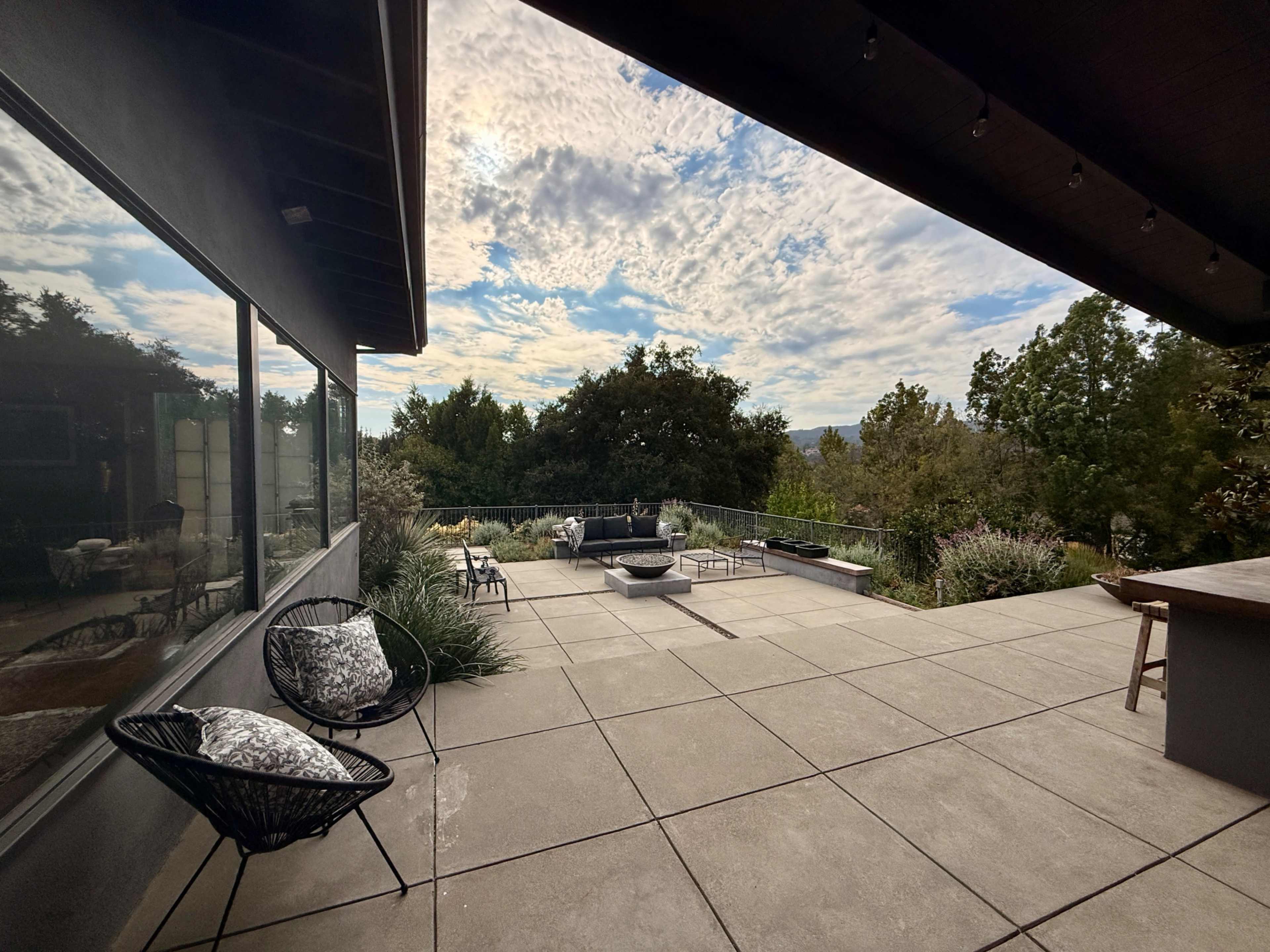 A spacious outdoor patio features modern seating, a fire pit, and views of surrounding trees and hills under a partly cloudy sky.