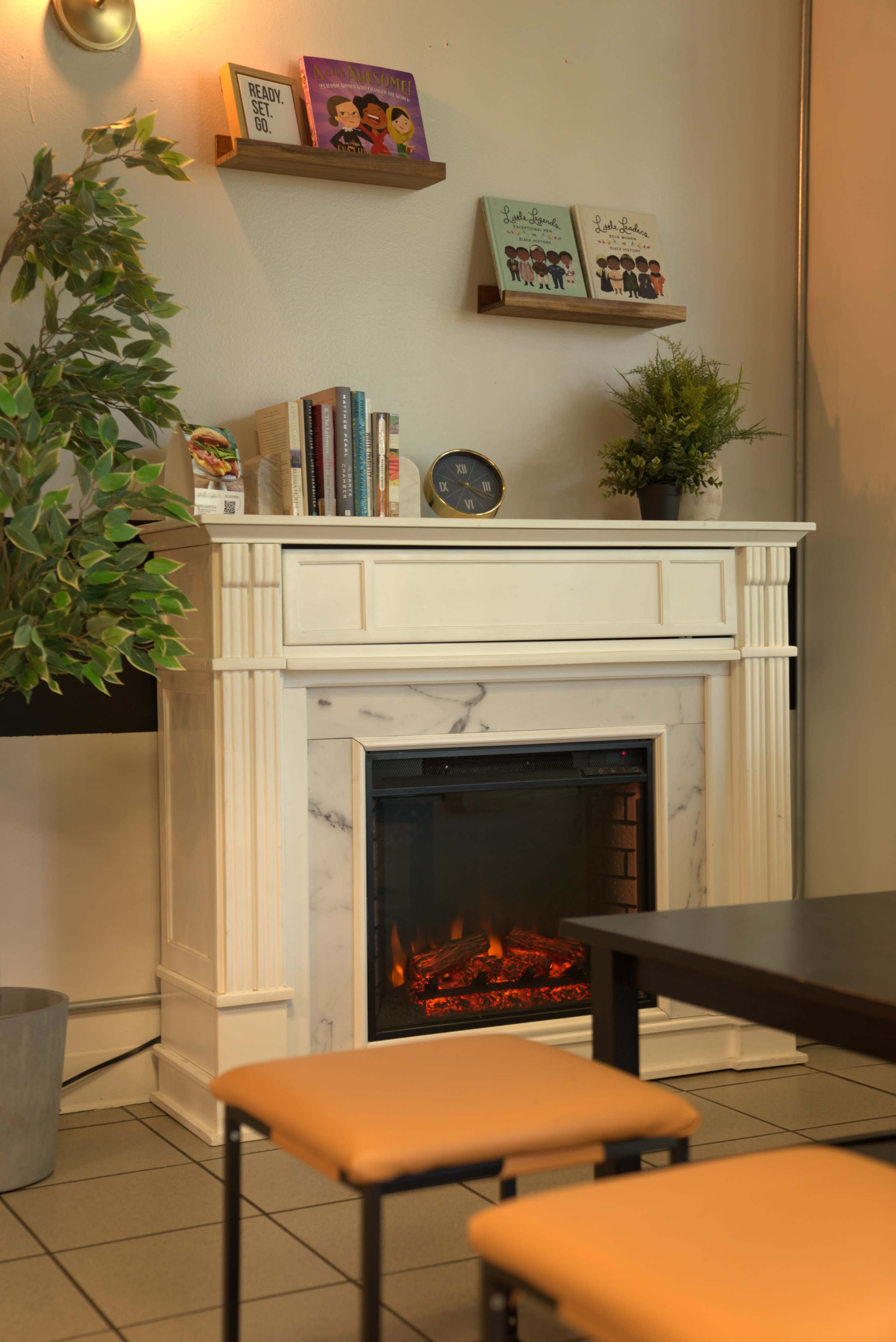 A modern fireplace with an artificial flame is set in a white mantel, accompanied by books and framed artwork on the wall above it, with a table and chairs in the foreground.