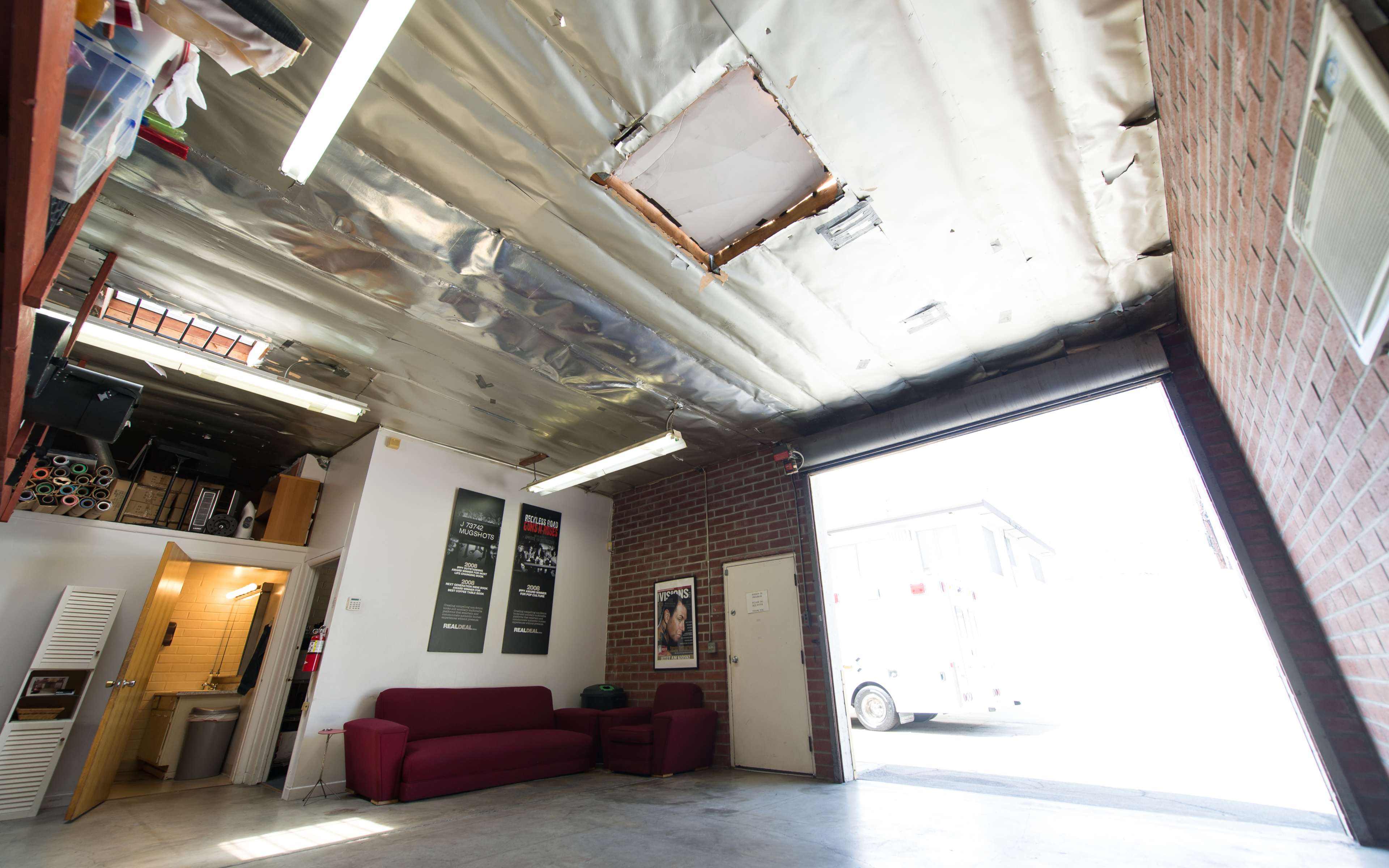 The image shows an unfinished garage space with a couch, a missing ceiling panel, and a large open door leading to an exterior area.