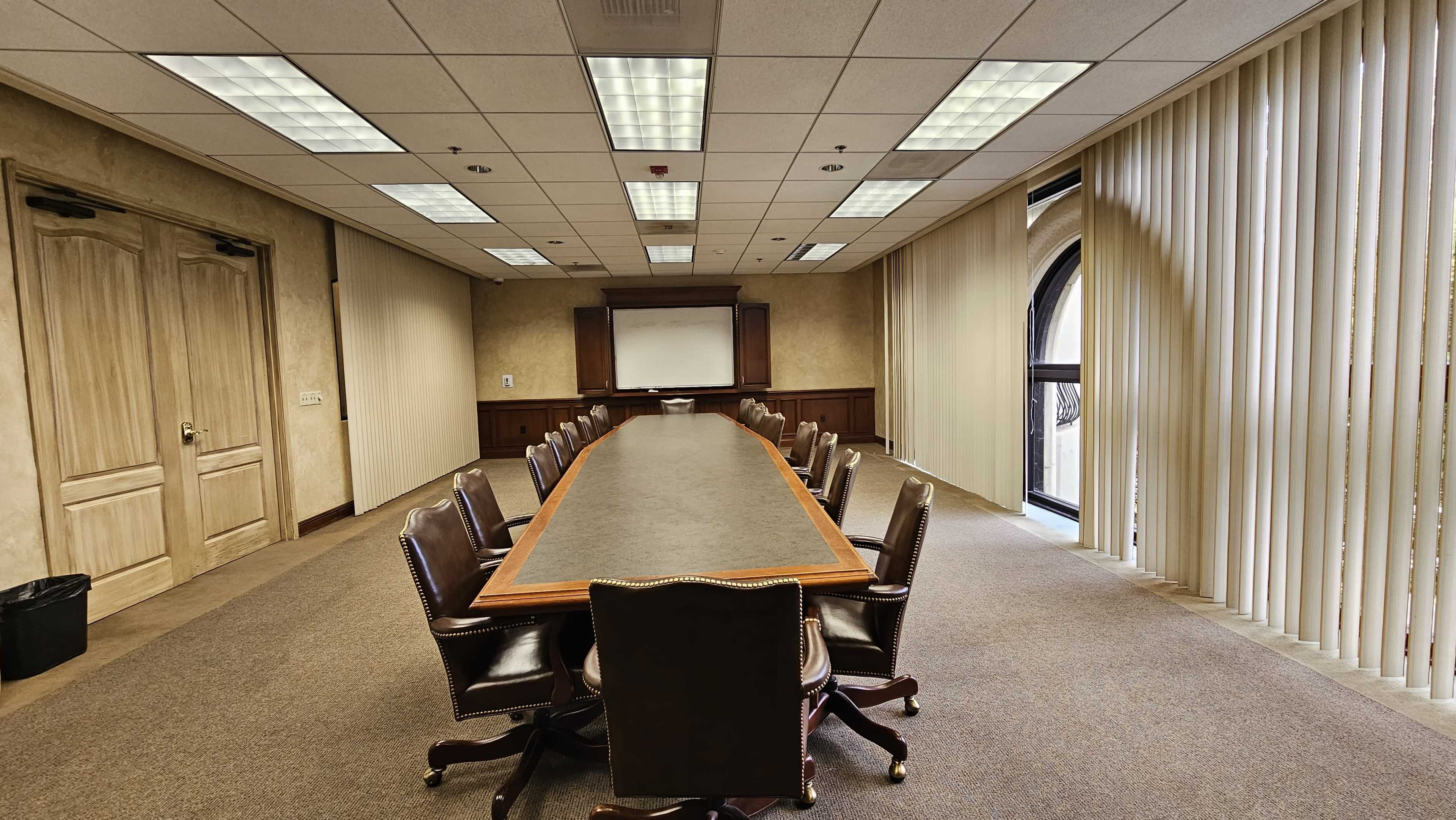 A long wooden conference table is surrounded by leather chairs in a well-lit meeting room with large windows and beige walls.