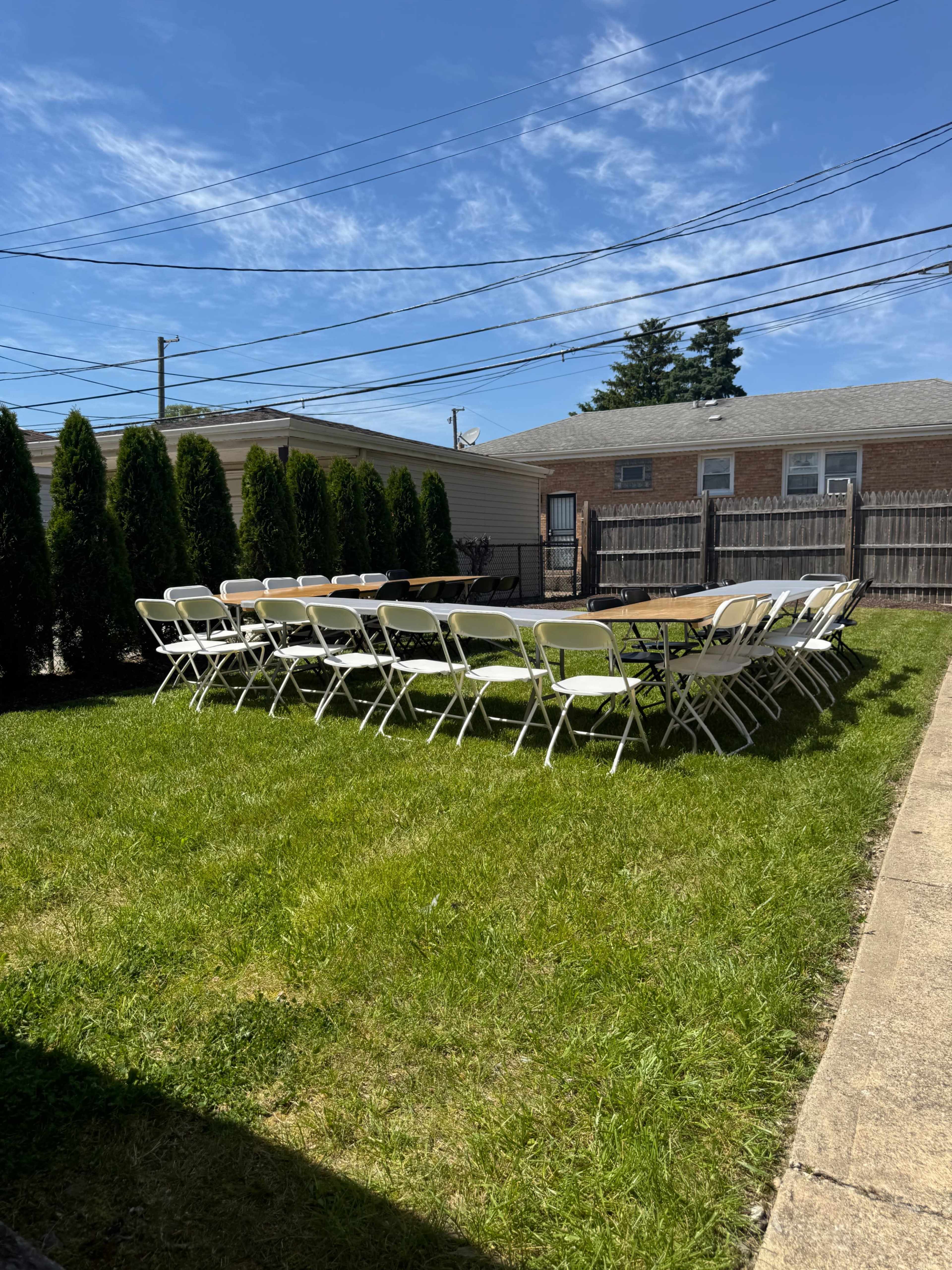 A row of white folding chairs is arranged around several wooden tables in a grassy backyard.