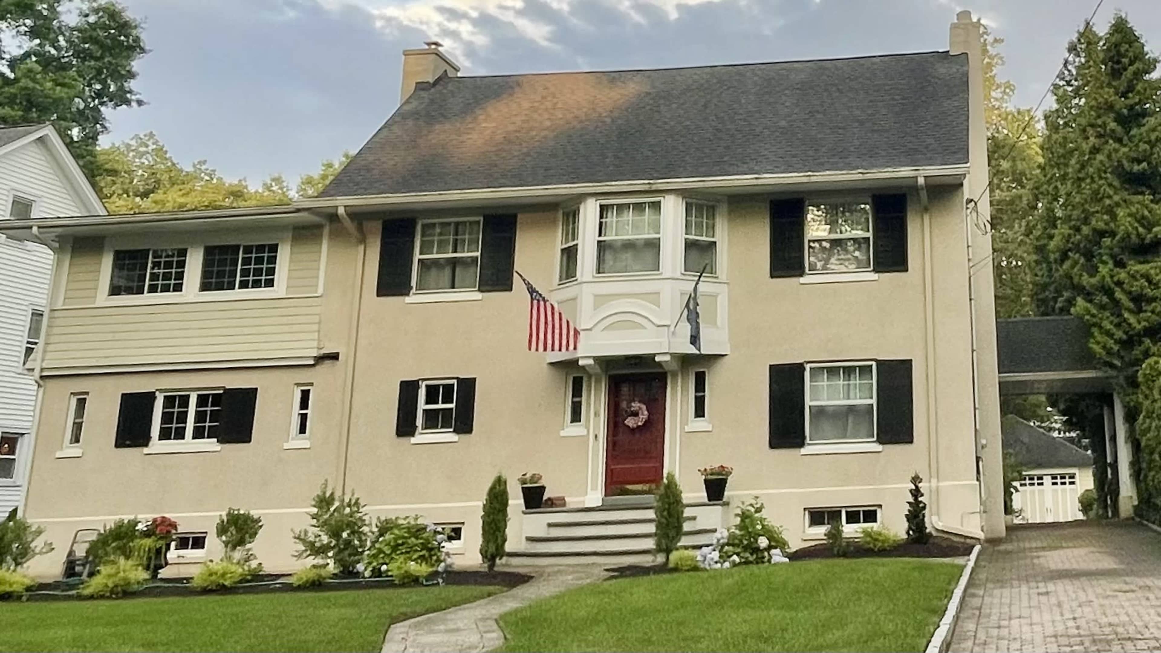 The image shows a two-story house with a central bay window, American flags hanging from the porch, and a well-maintained front yard.